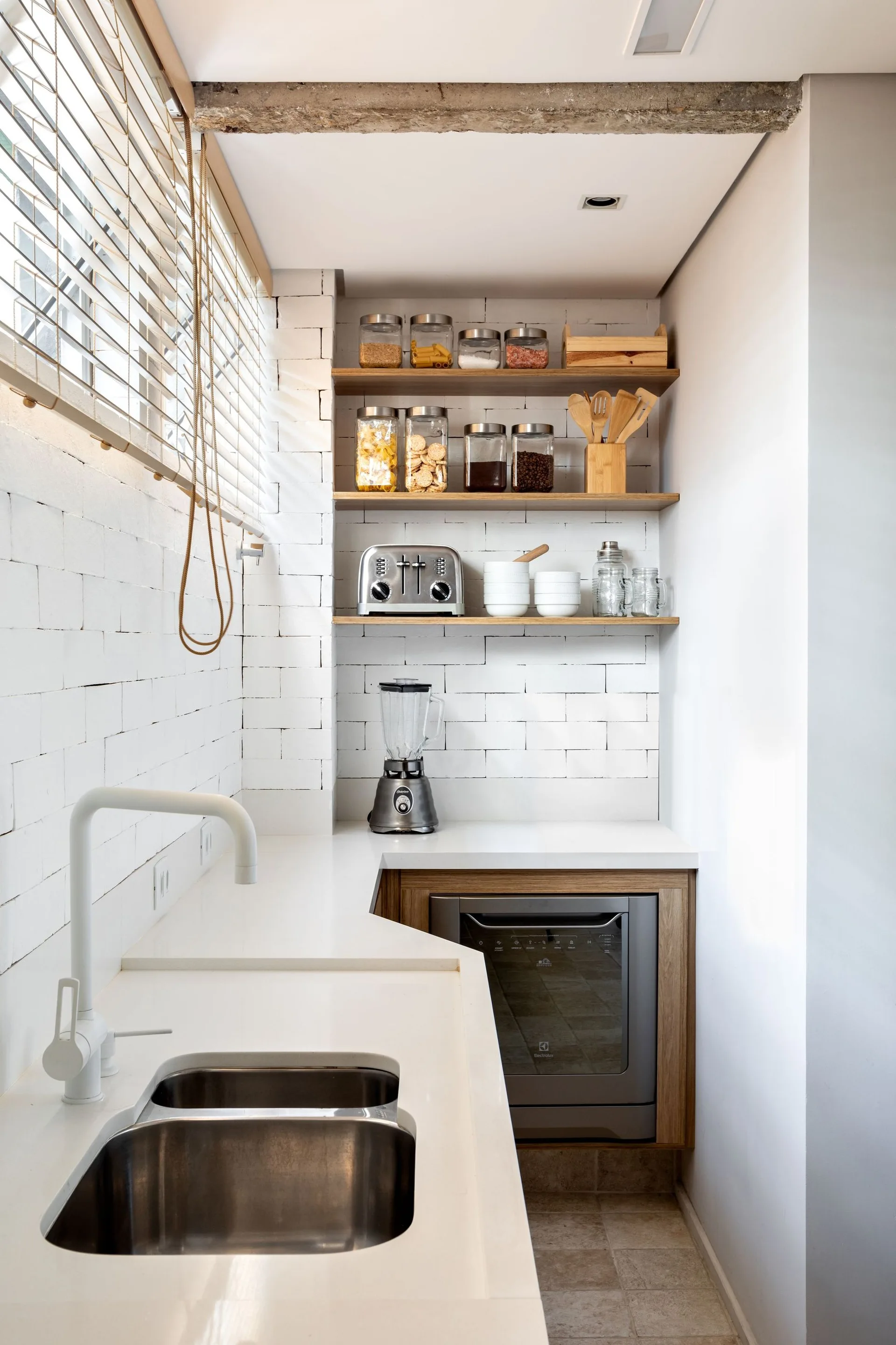 A compact kitchen featuring a white countertop with an integrated sink, white painted brick walls, and three open wood shelves holding glass storage jars.