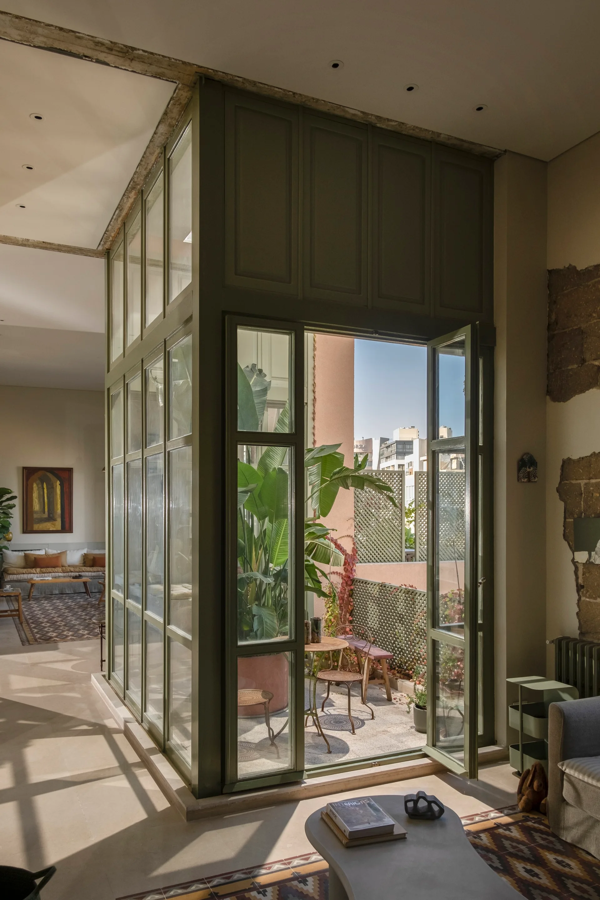 An olive green glass enclosure with double doors leads from a light-filled living room to a sunny outdoor terrace.