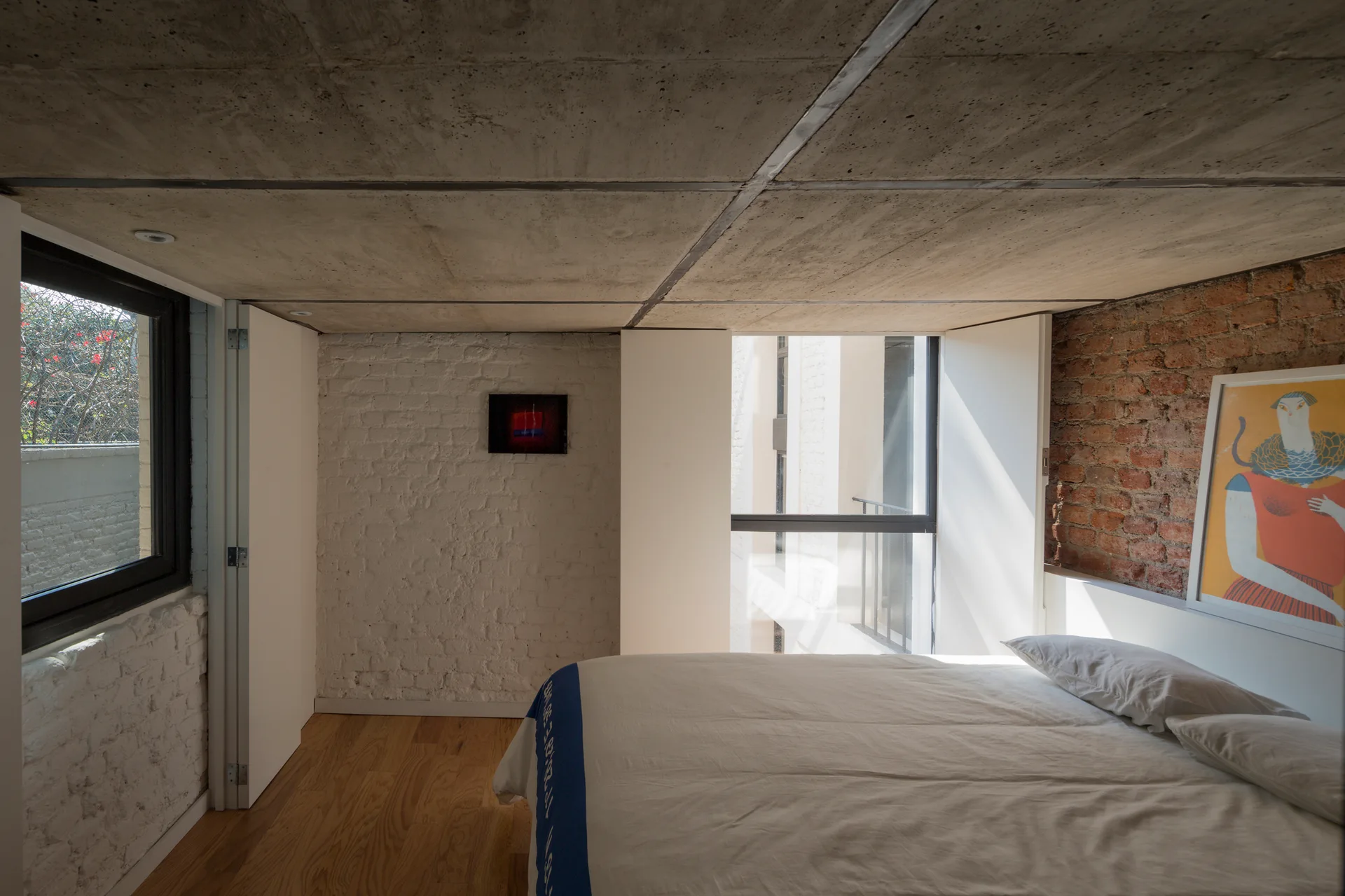 A modern minimalist loft bedroom featuring a low raw concrete ceiling, a wood floor, a bed with white sheets, and walls of painted white and red brick.