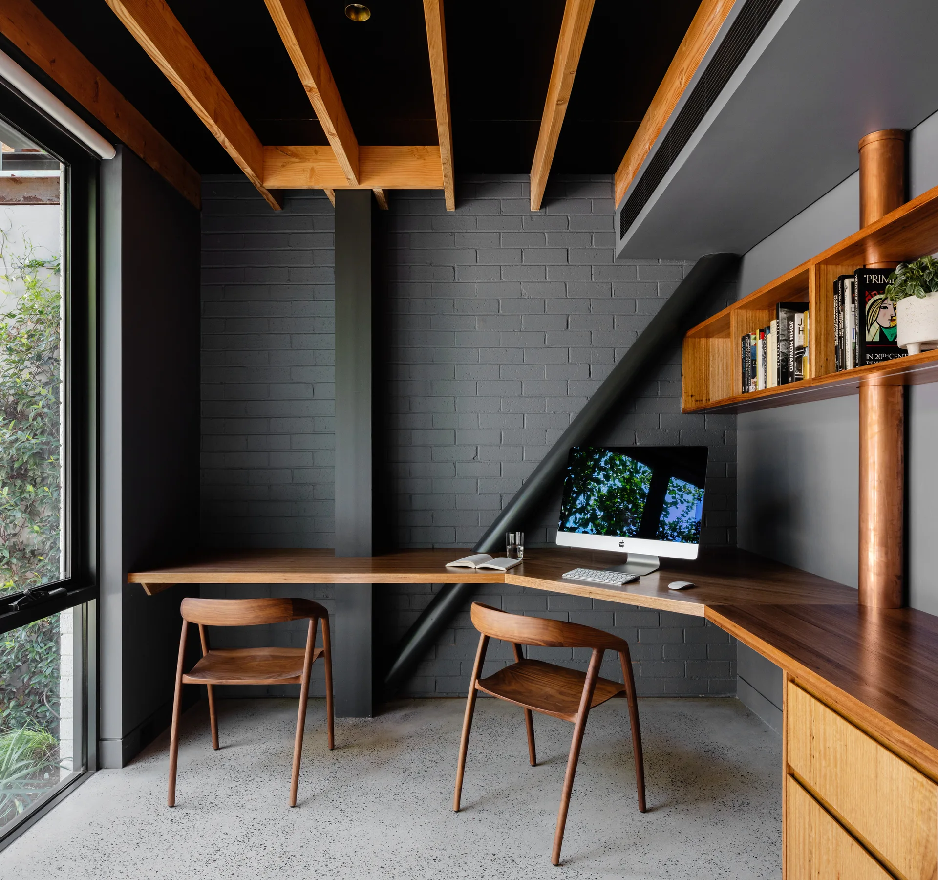 A modern home office featuring a dark gray painted brick wall, a custom wooden corner desk with matching chairs, and exposed ceiling beams.