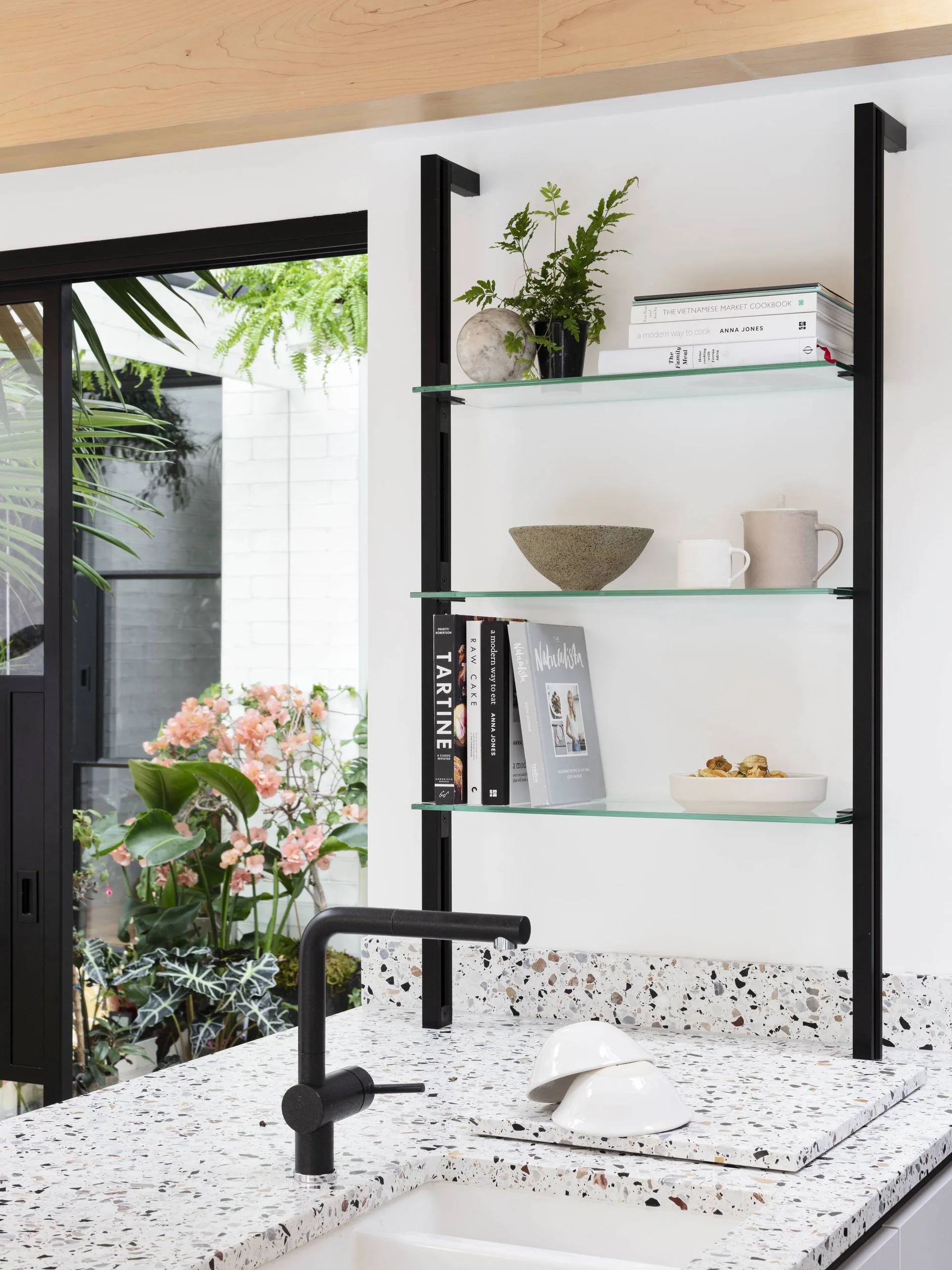 A white terrazzo kitchen countertop with a black faucet next to wall-mounted glass shelving holding books and plants, with a patio visible outside.