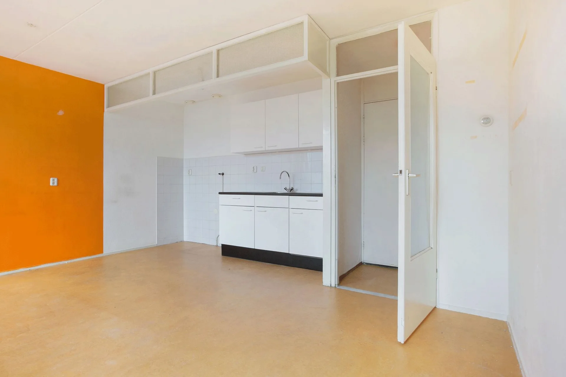 A small white kitchenette features a square tile backsplash, a vibrant orange accent wall, yellow flooring, and an open glass-paneled door.