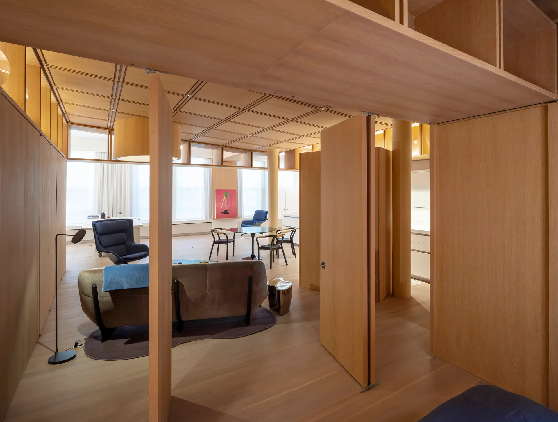 An open-concept living space features extensive light wood wall paneling, a wooden grid ceiling, modern minimalist furniture, and large windows.