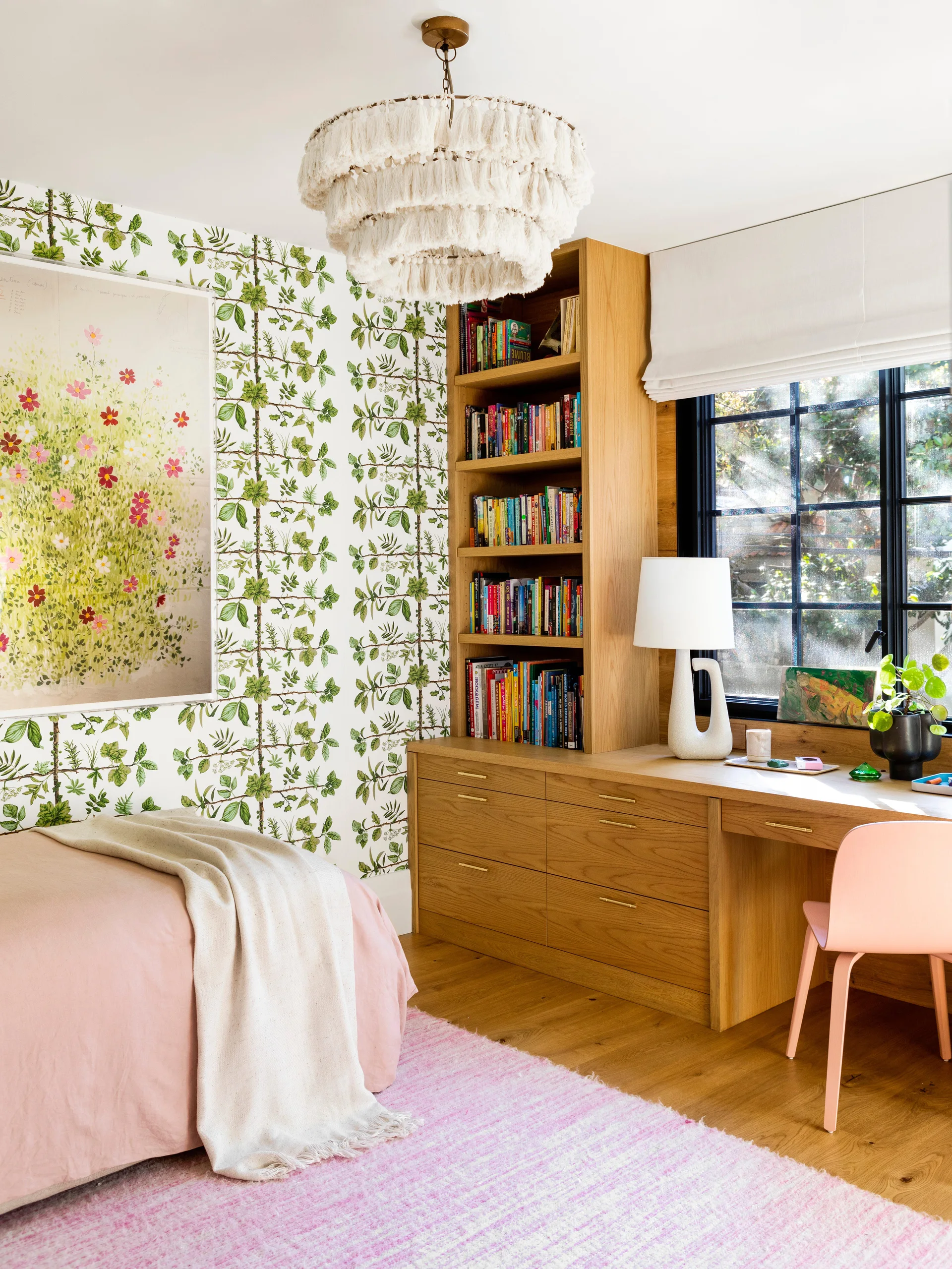 A bedroom features green botanical wallpaper, a pink bed, and a built-in oak desk with an integrated bookshelf next to a black-framed window.