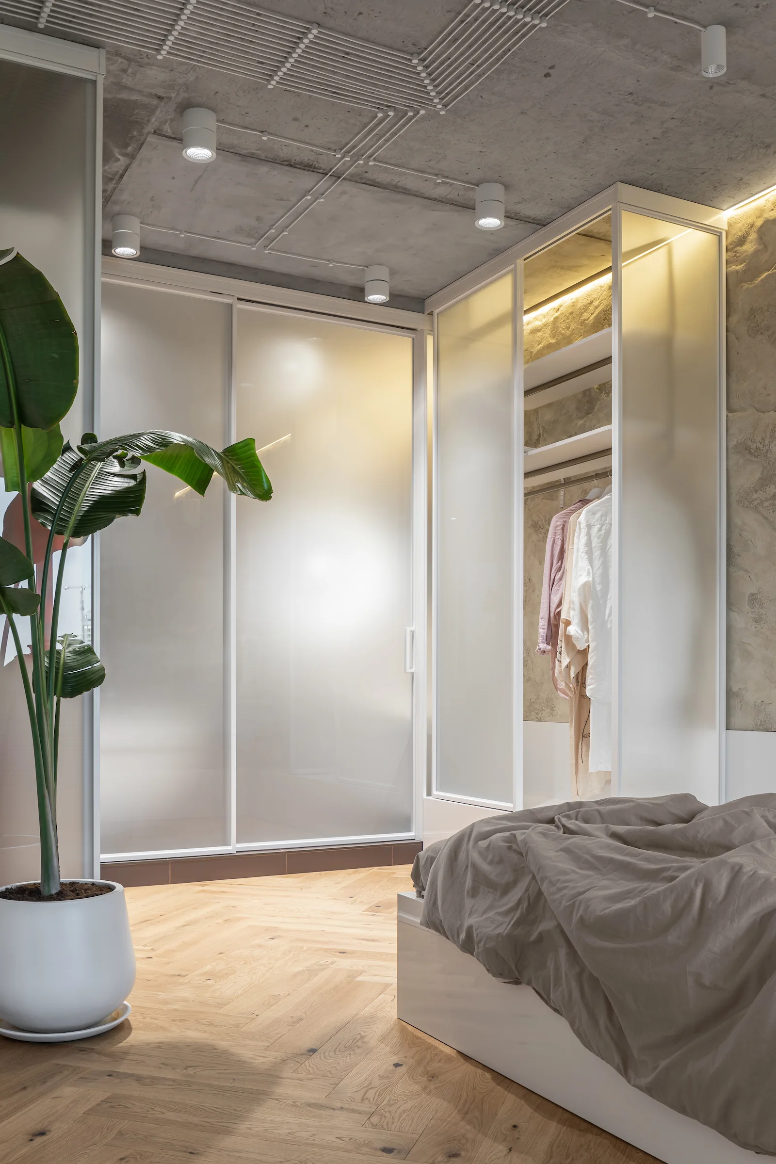 A modern bedroom features a frosted glass sliding door wardrobe, an exposed concrete ceiling with spotlight fixtures, and herringbone hardwood flooring.