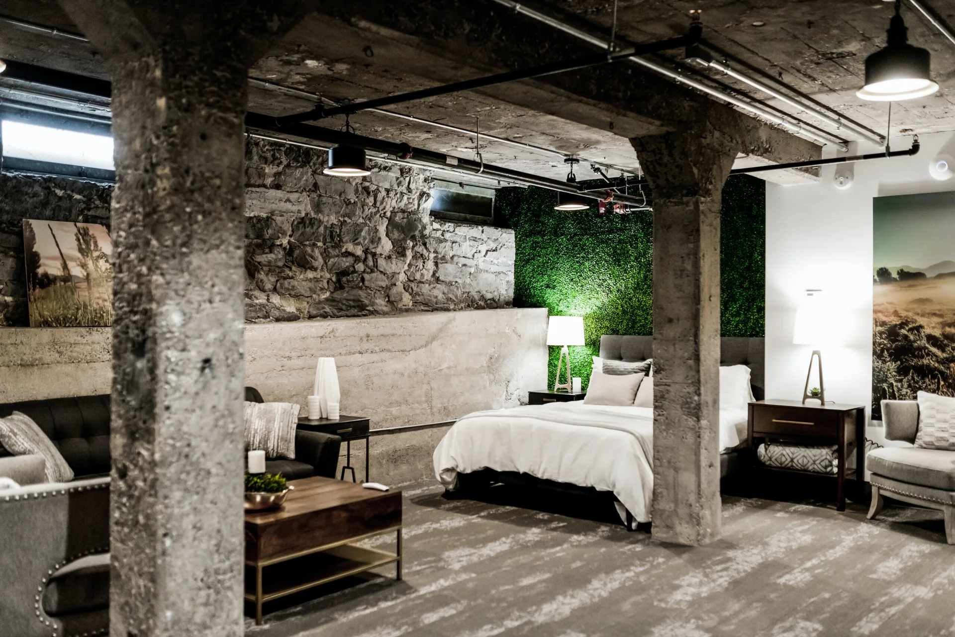An industrial basement bedroom featuring raw concrete pillars, an exposed stone wall, a green foliage panel behind the bed, and warm table lamps.