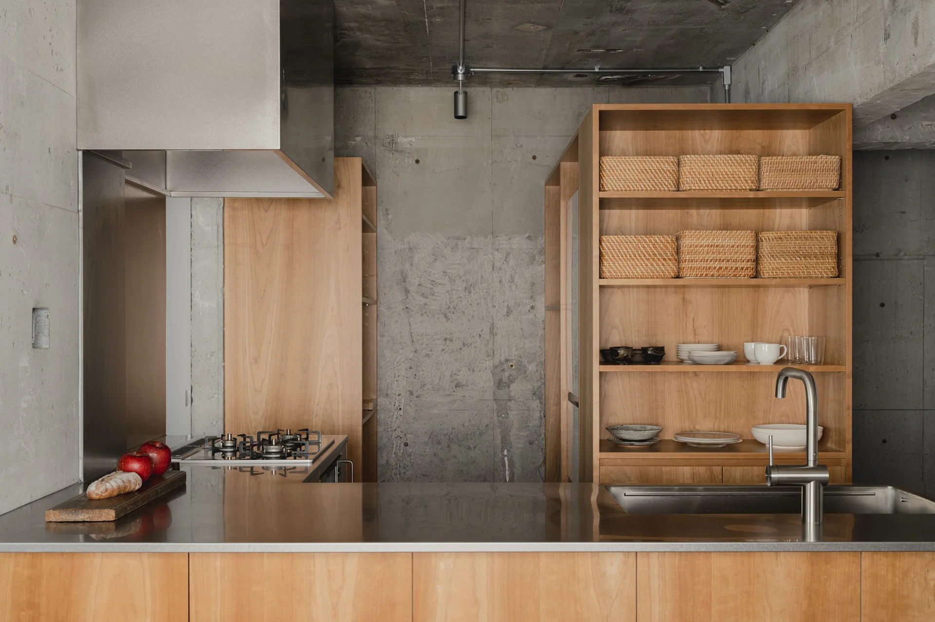 A minimalist industrial kitchen features a stainless steel countertop, warm wood storage shelving with woven baskets, and raw concrete walls.