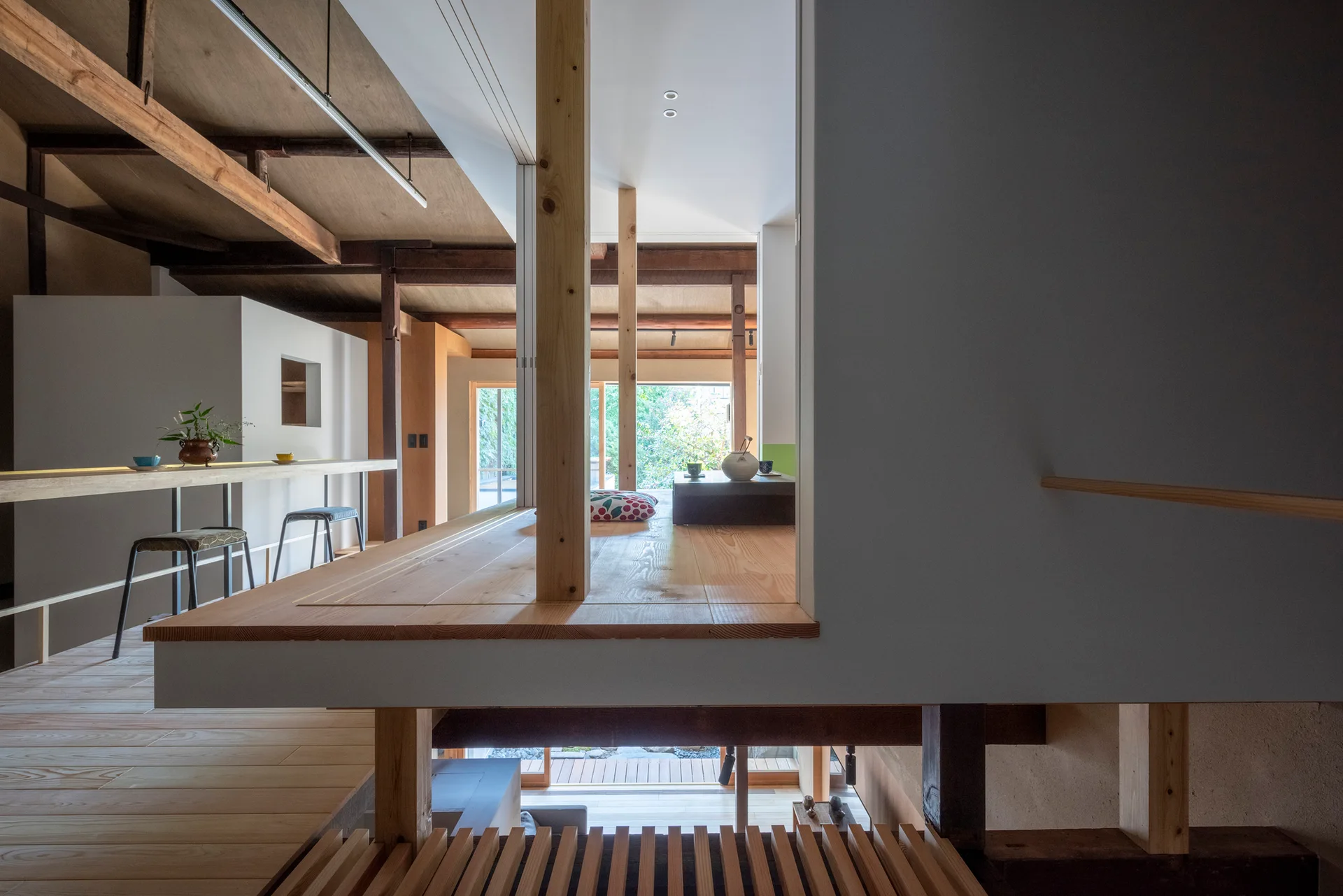 A minimalist Japanese interior showcases an elevated wooden platform, exposed ceiling rafters, vertical timber posts, and a bar counter with stools.