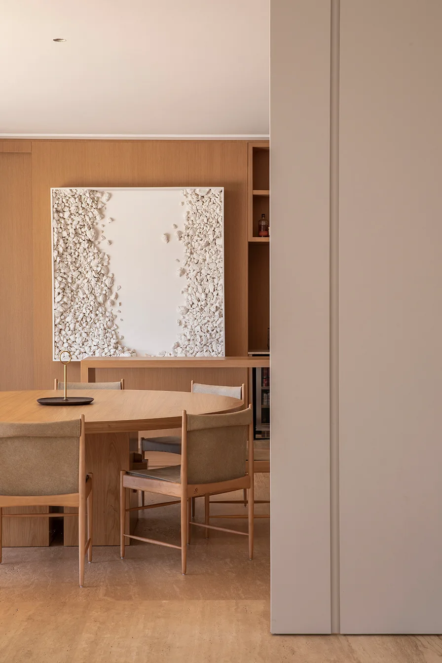 A minimalist dining room features a round wood table and chairs in front of a wood-paneled wall with a large textured white sculpture.