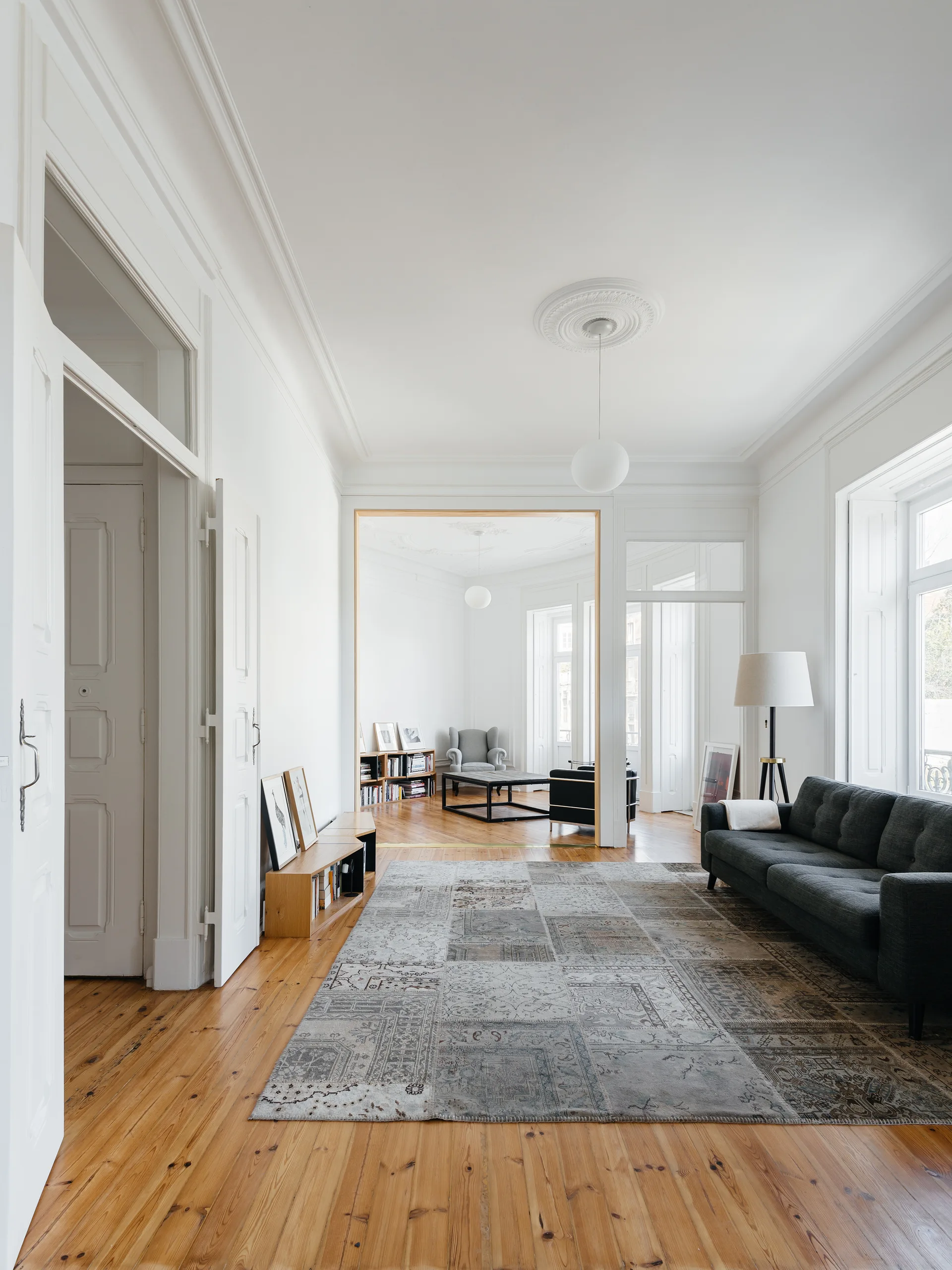 A spacious white living room features a dark grey sofa, a faded patchwork rug on wood floors, and historic ceiling trim.