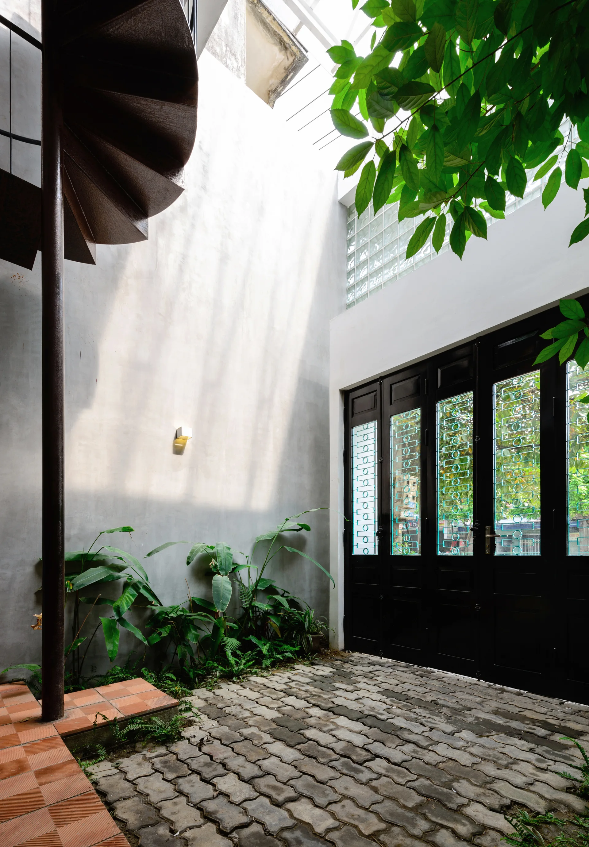 A semi-open concrete courtyard features a metal spiral staircase, textured stone pavers, lush green plants, and black-framed glass doors.