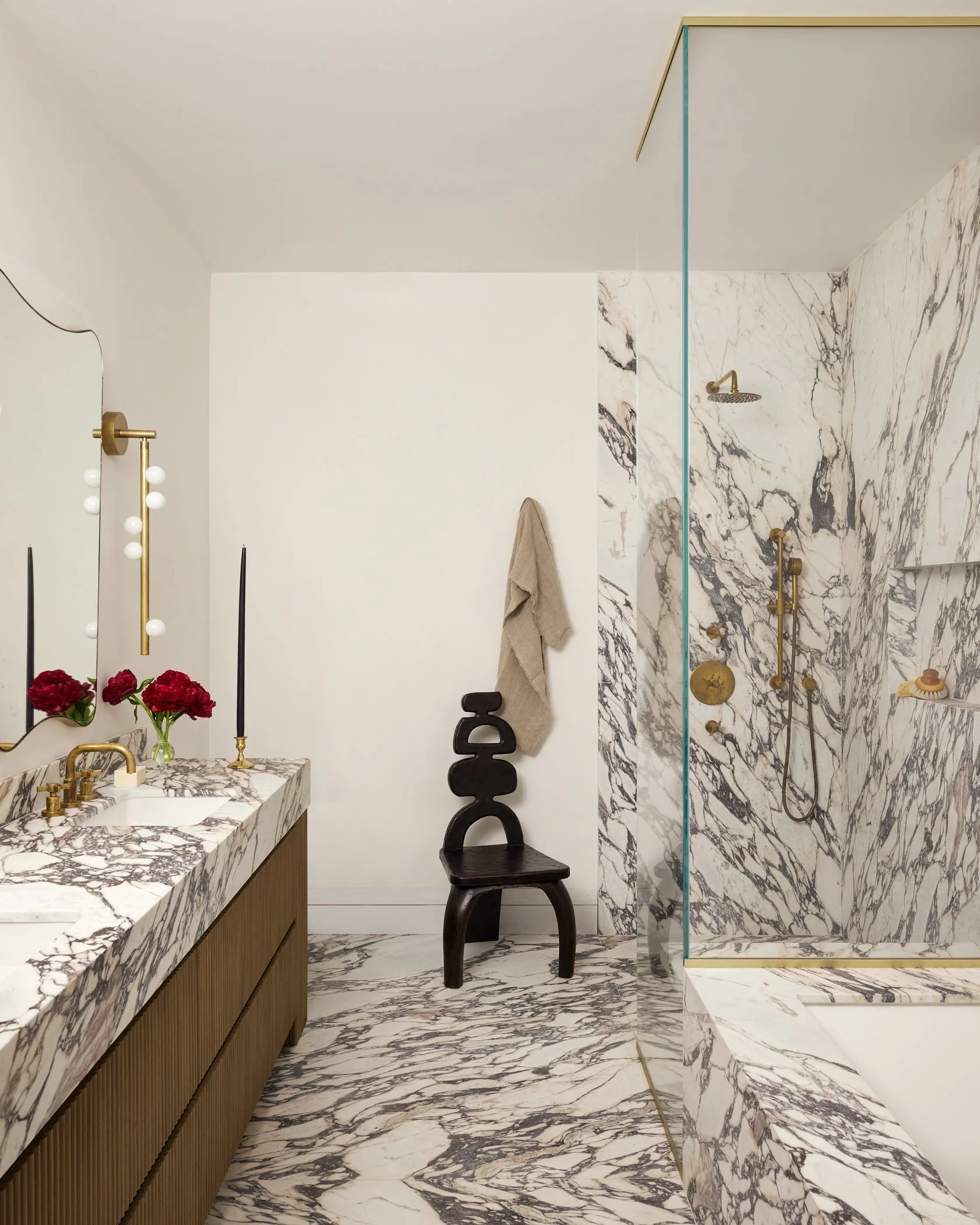 A contemporary bathroom with heavily veined white and grey marble surfaces, a fluted wood vanity with brass fixtures, and a sculptural dark stool.