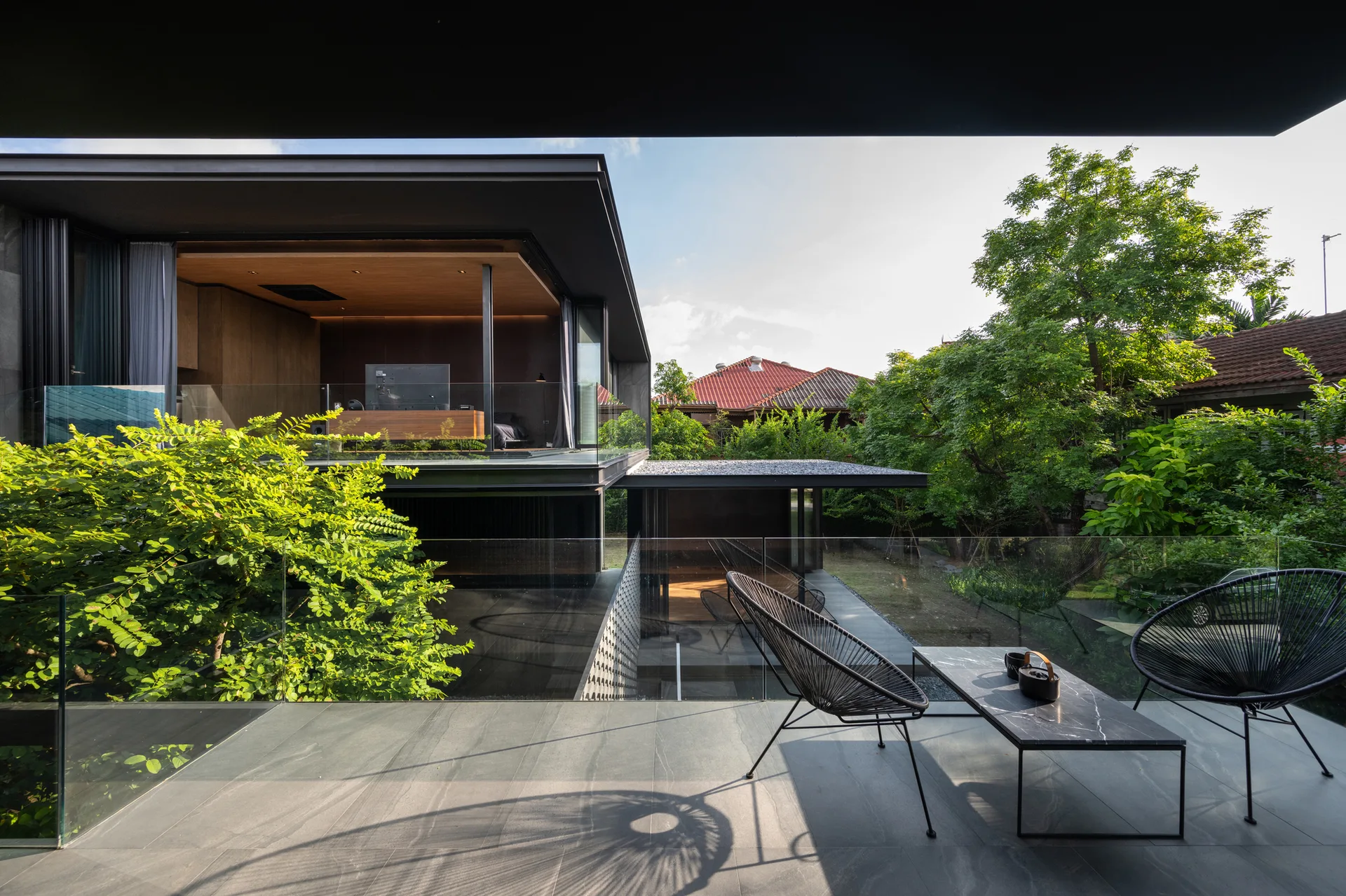 A modern stone-tiled terrace features two black wire chairs and a low coffee table overlooking a lush garden through a clear glass railing.