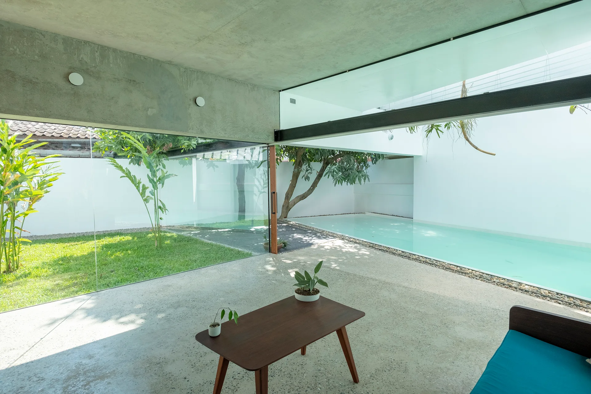 A minimalist living space features an exposed concrete ceiling, a low wooden coffee table, and floor-to-ceiling glass walls looking out to a pool.