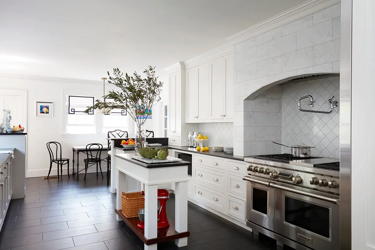 A bright white kitchen with a professional stainless steel range under a marble alcove, a central island, and dark tiled flooring.