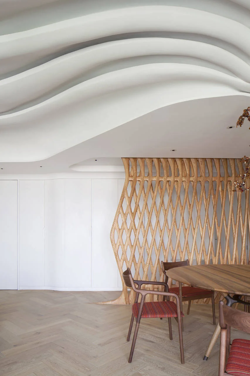 A modern dining area features a circular wooden table, chairs with red seats, a sculptural lattice wood screen, and a wavy layered ceiling.