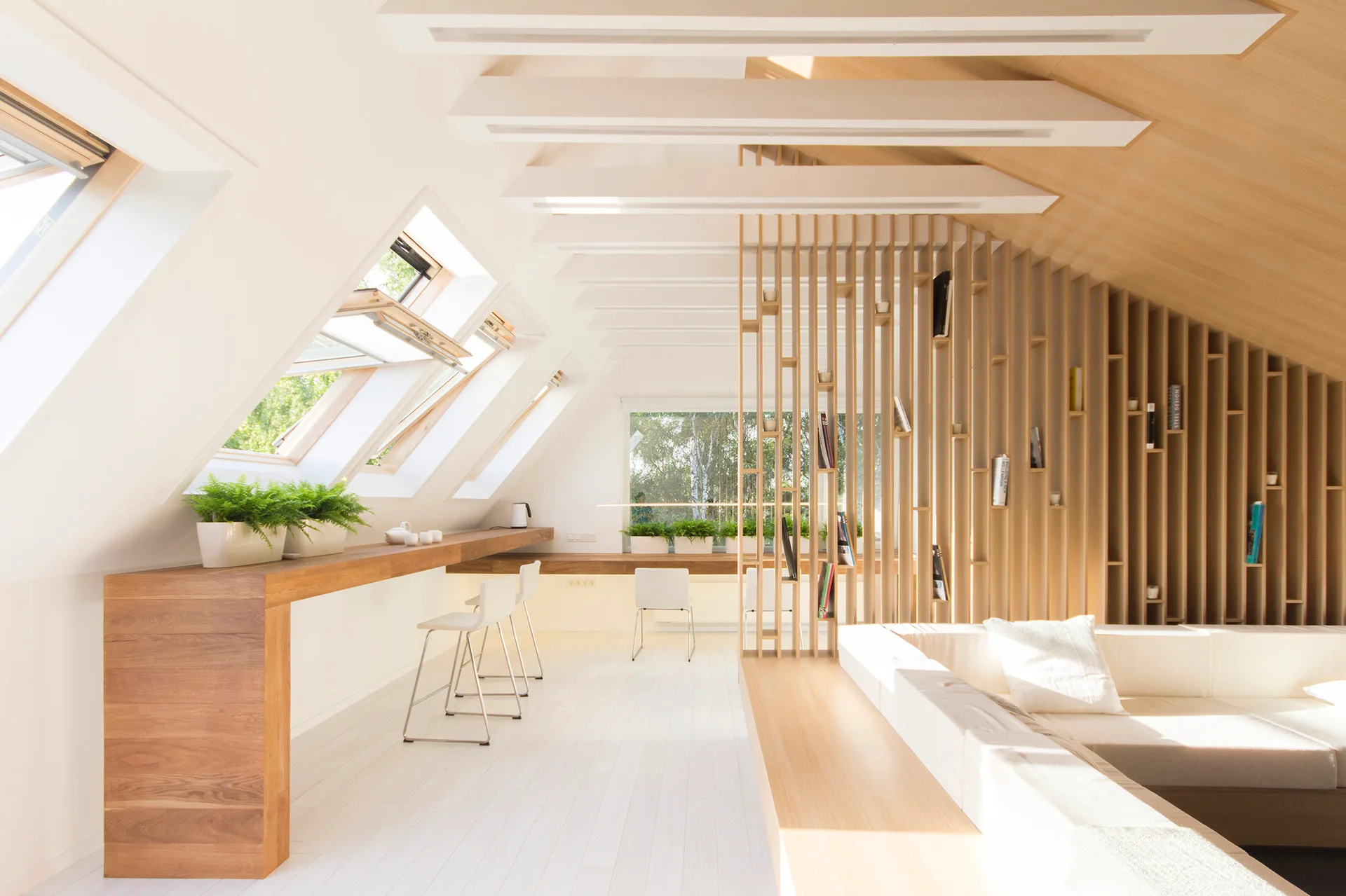 A bright attic loft features a long wood desk under skylights, a vertical timber slat divider, and white minimalist seating.
