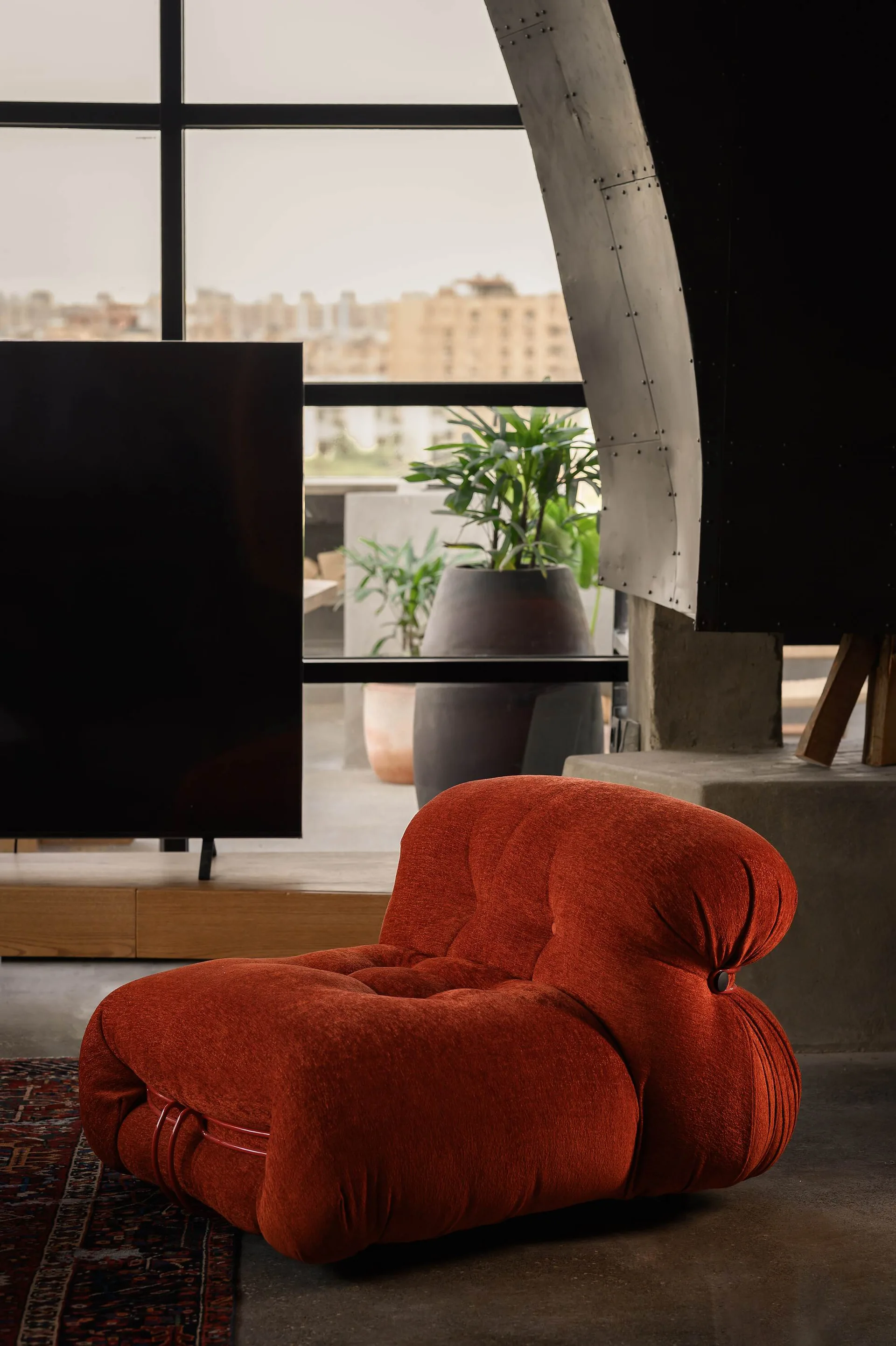 A tufted rust-red lounge chair sits on a concrete floor in front of a large black-framed window overlooking an outdoor terrace.