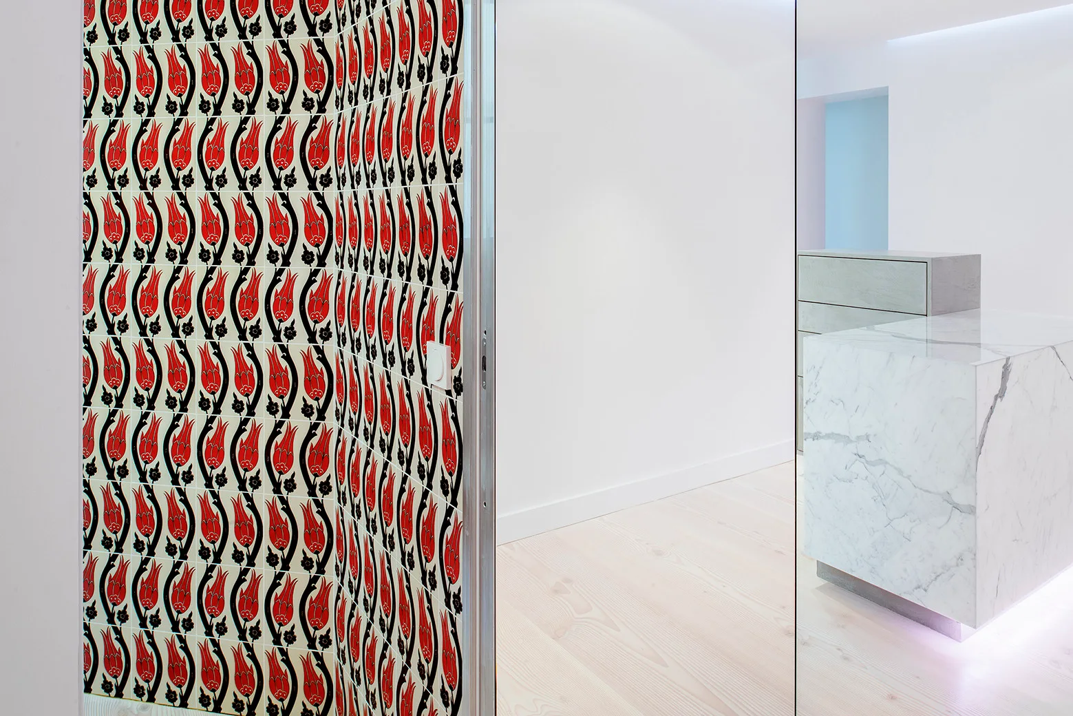 A wall tiled in a red and black tulip pattern stands adjacent to a modern white kitchen featuring a marble island with integrated lighting.