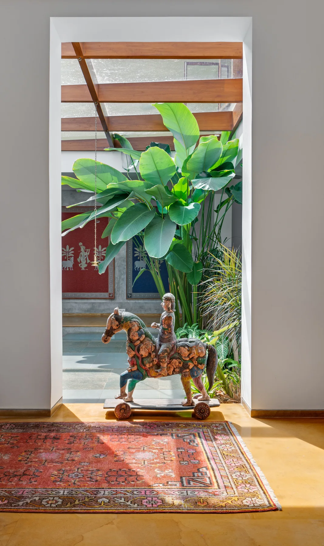 A white doorway frames an indoor courtyard with a painted wooden horse sculpture, lush green plants, and a wood-beamed glass skylight.