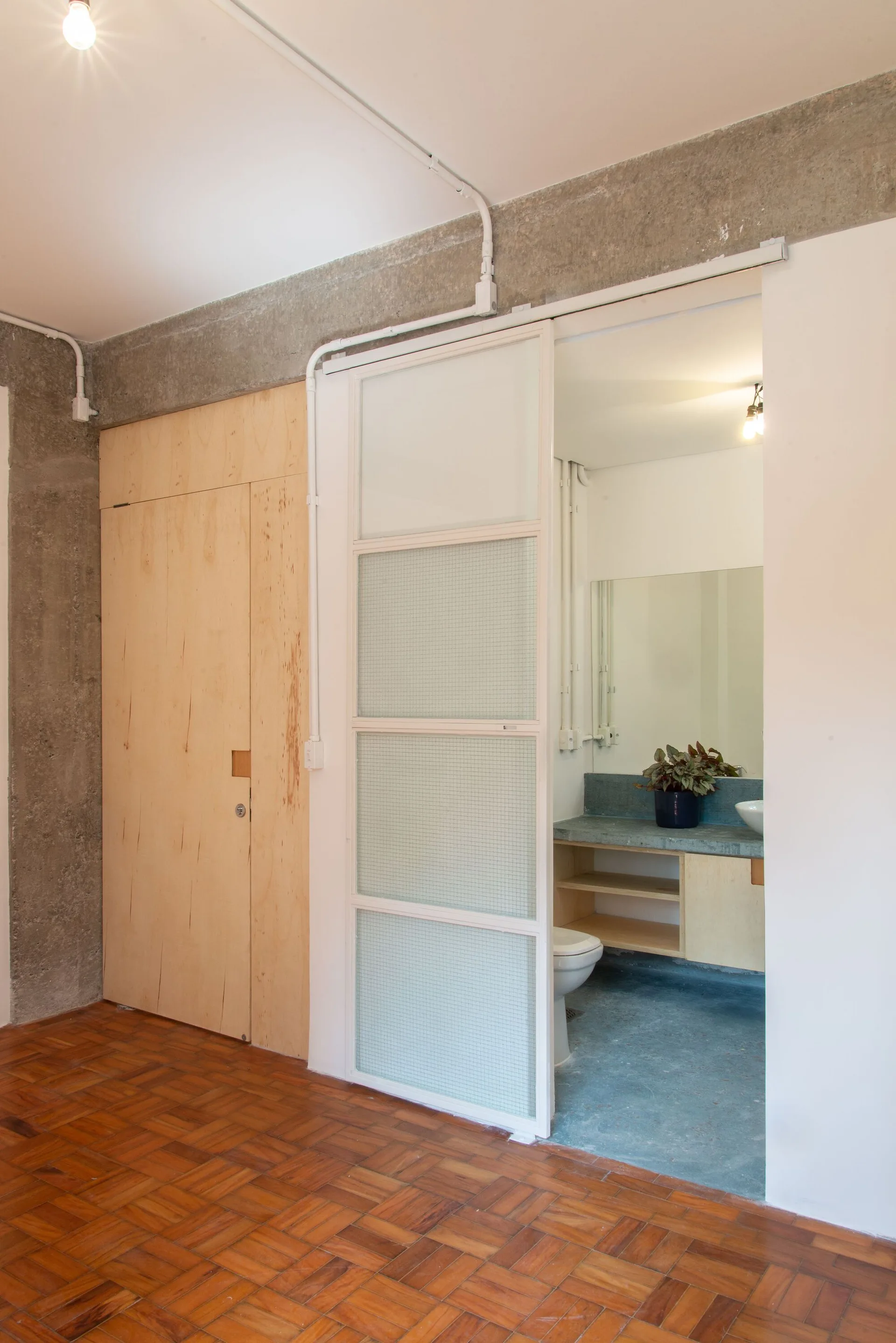 A white sliding door with grid glass panels opens to an industrial bathroom featuring a concrete vanity and blue floor next to a parquet wood hallway.