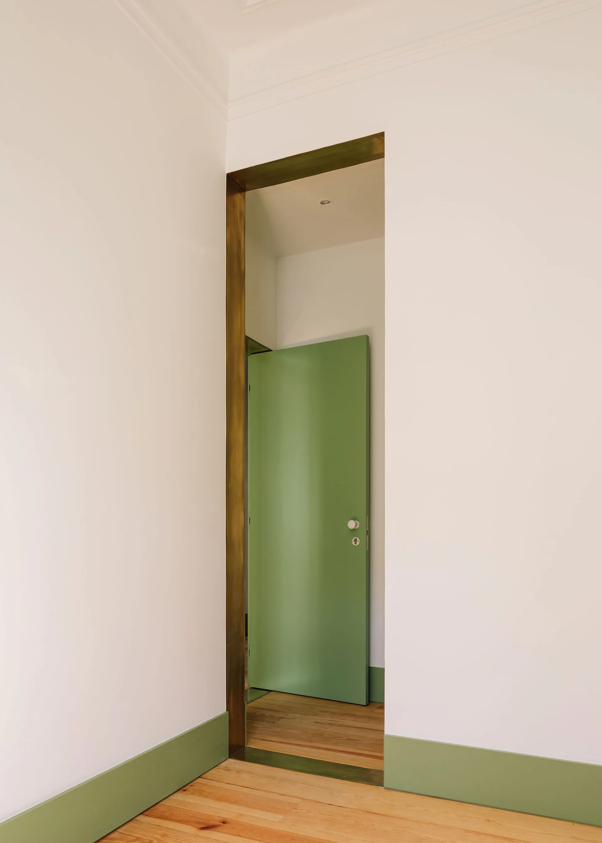 A minimalist hallway doorway wrapped in a brass metal frame features a sage green door and matching green baseboards against white walls.