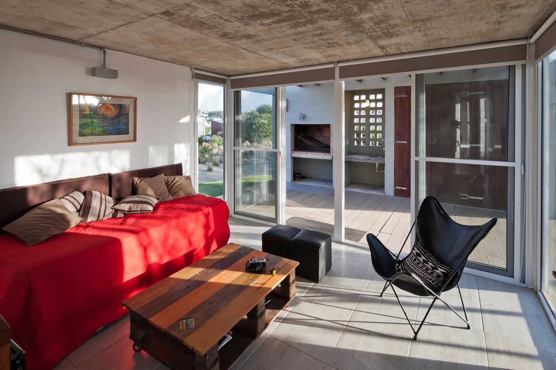 A modern living room featuring a bright red sofa, a wooden coffee table, and a black butterfly chair underneath an exposed concrete ceiling.