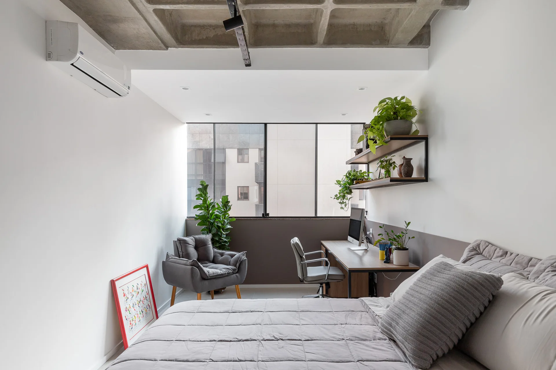 A modern bedroom and home office featuring a grey quilted bed, a wooden desk under floating shelves with plants, and an exposed concrete waffle ceiling.