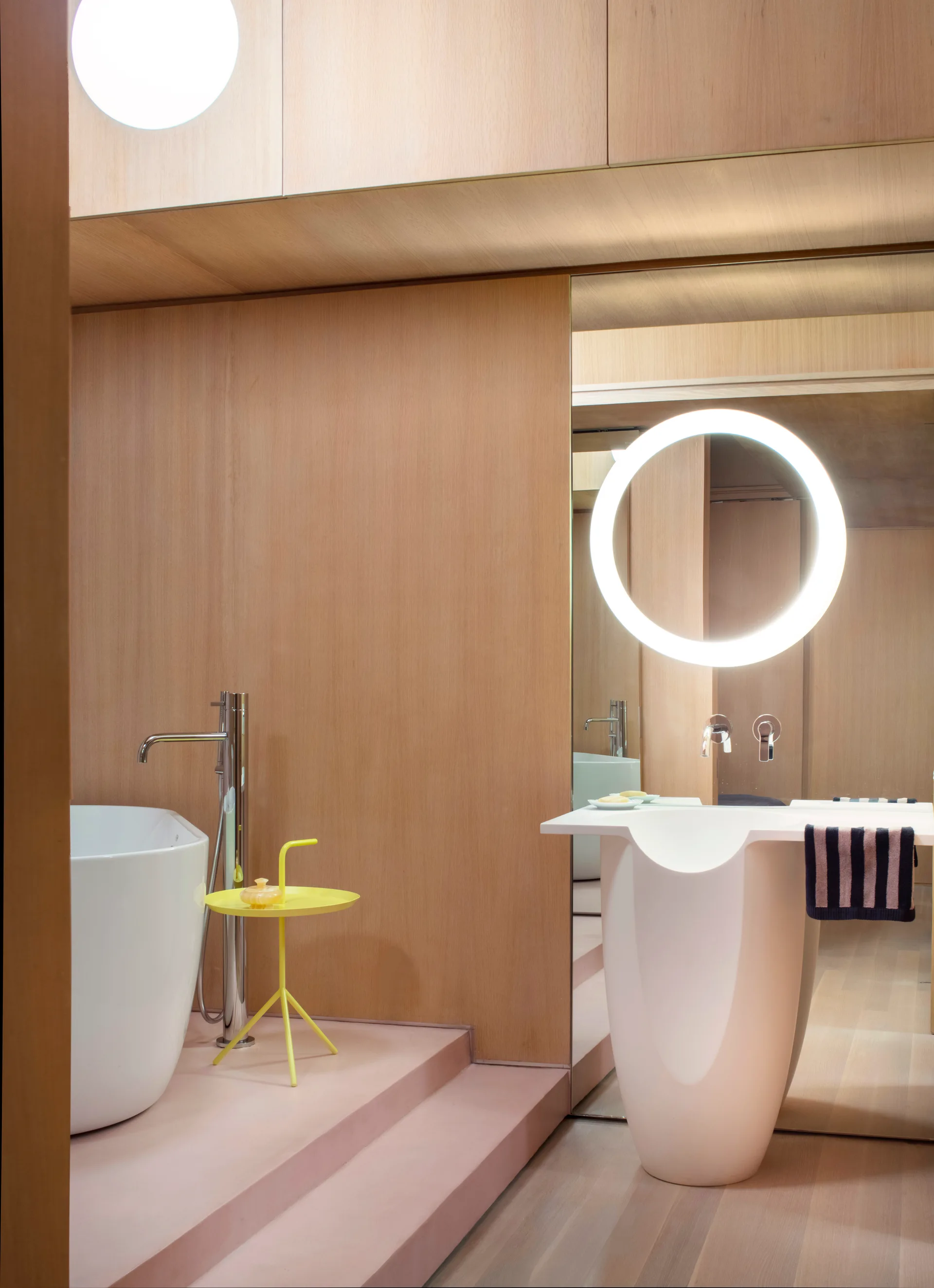A bathroom features wood wall paneling, a raised pink platform with a soaking tub, and a sculptural pedestal sink beside an illuminated round mirror.