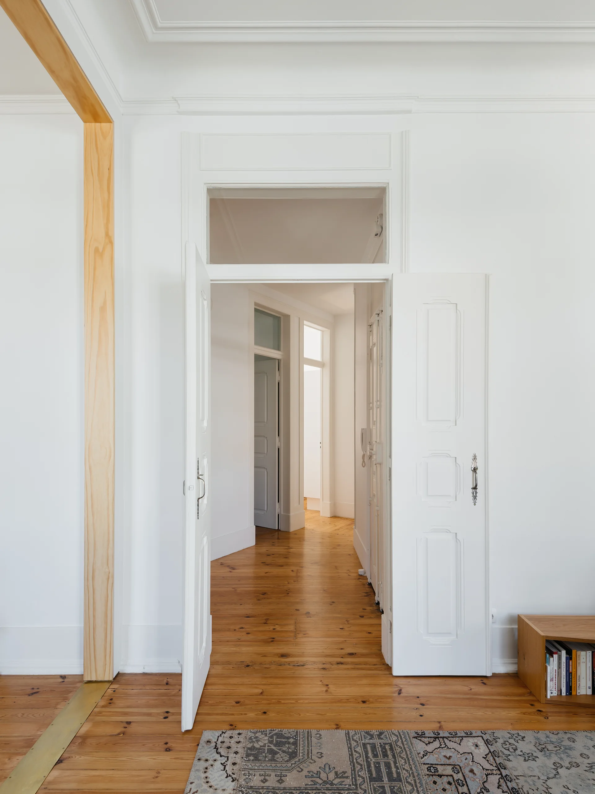White paneled double doors open into a bright hallway with warm pine floors, framed by a natural wood post and a patterned rug in the foreground.