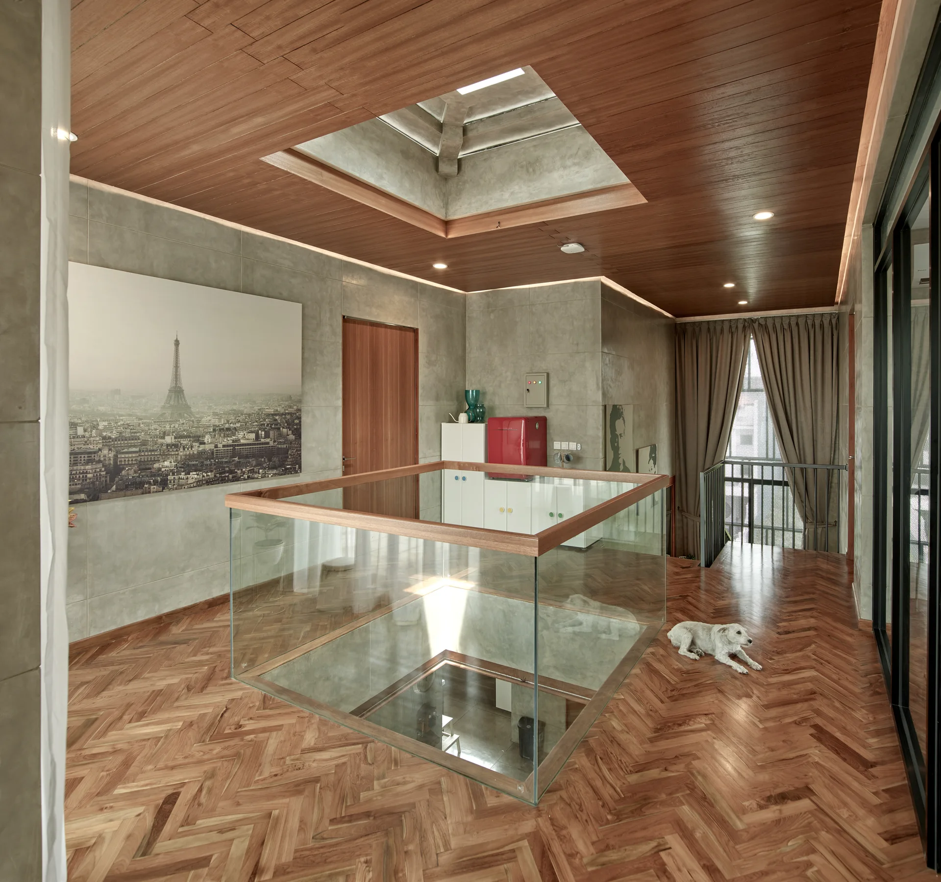 A modern mezzanine landing features a glass railing with wood trim, a warm wood plank ceiling with a skylight, and herringbone flooring.
