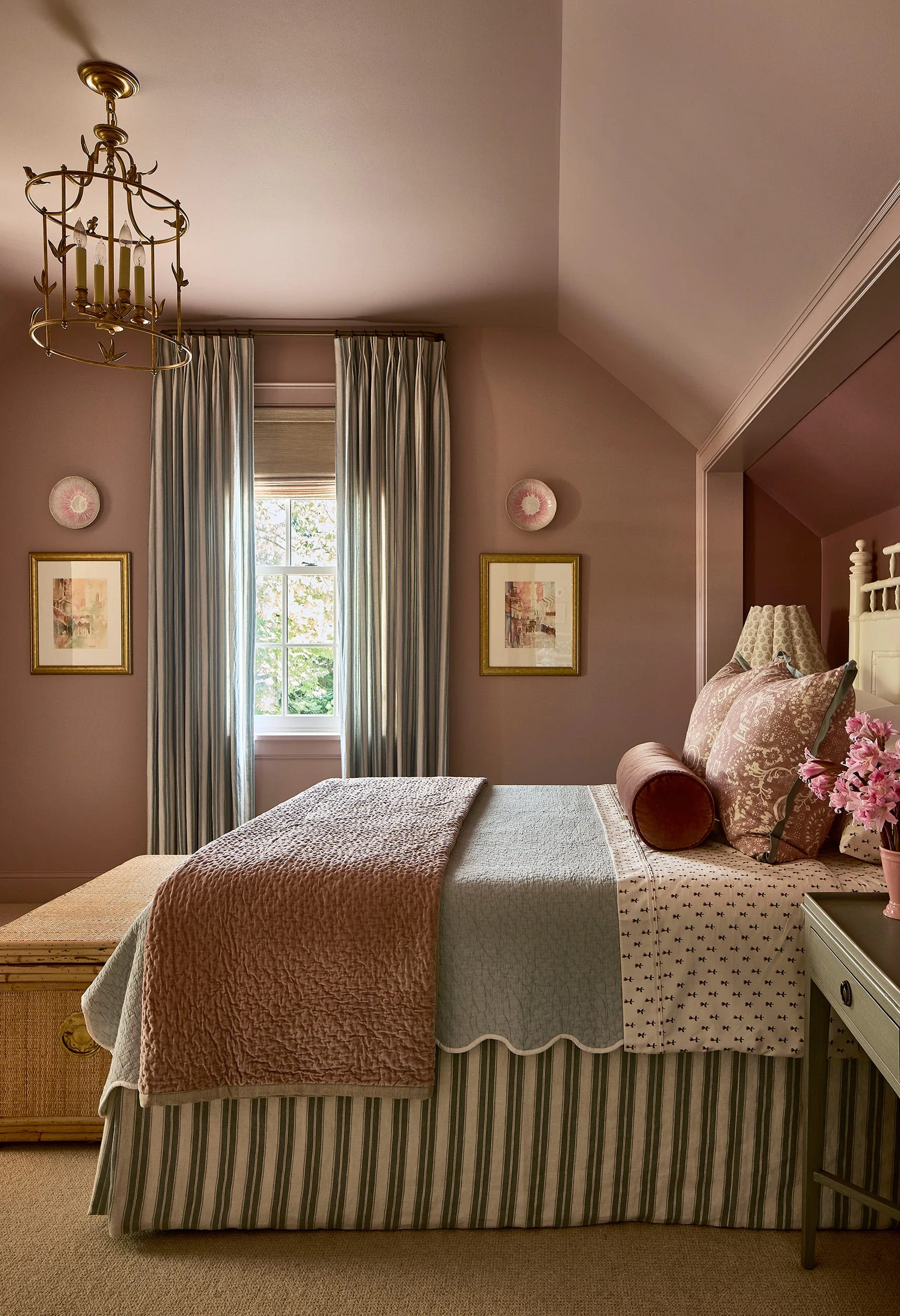 A bedroom with mauve walls and ceiling features a bed with layered patterned bedding, striped curtains framing a window, and a brass chandelier.