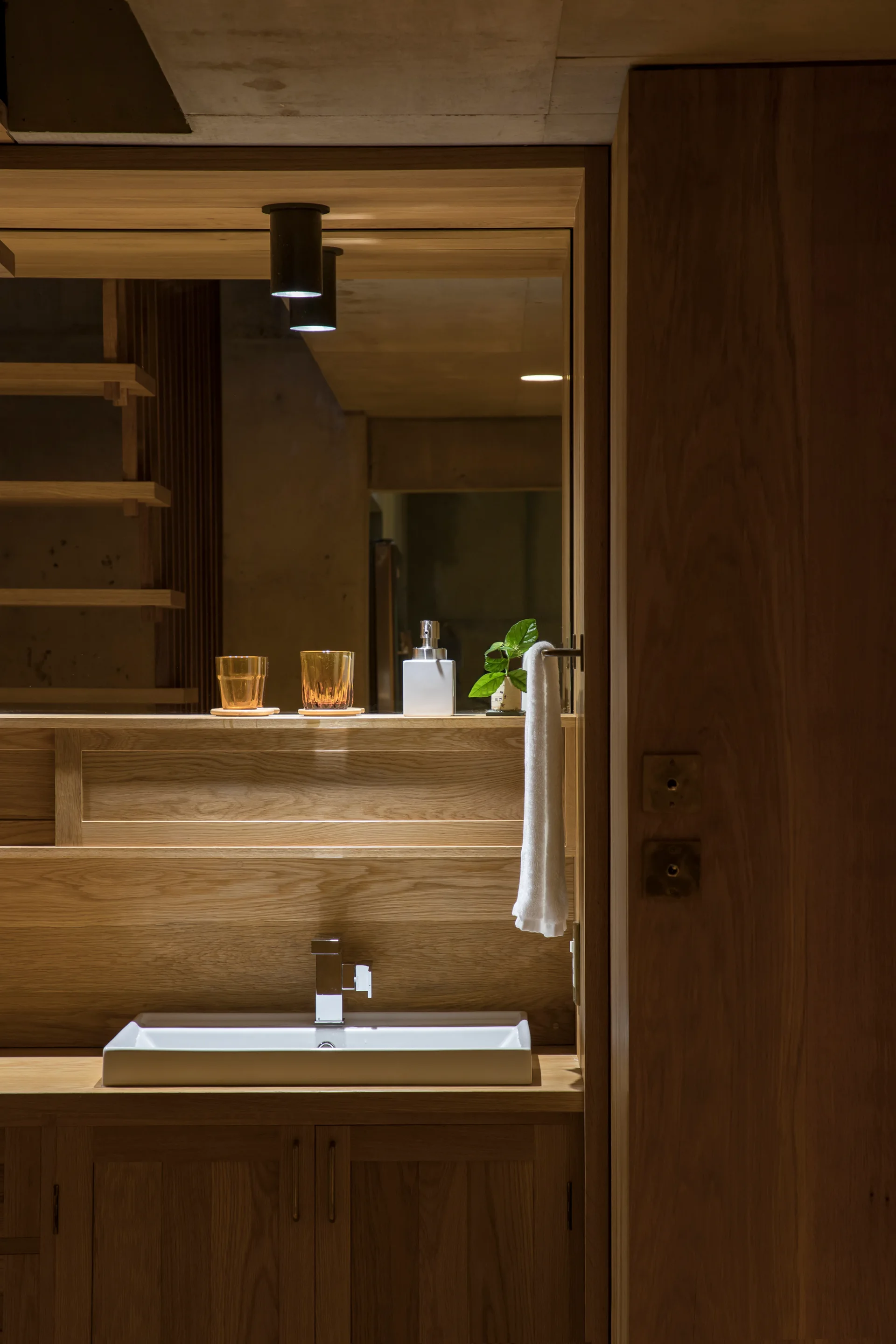 A minimalist bathroom vanity crafted from warm wood features a white rectangular sink, a large wood-framed mirror, and a small hanging hand towel.