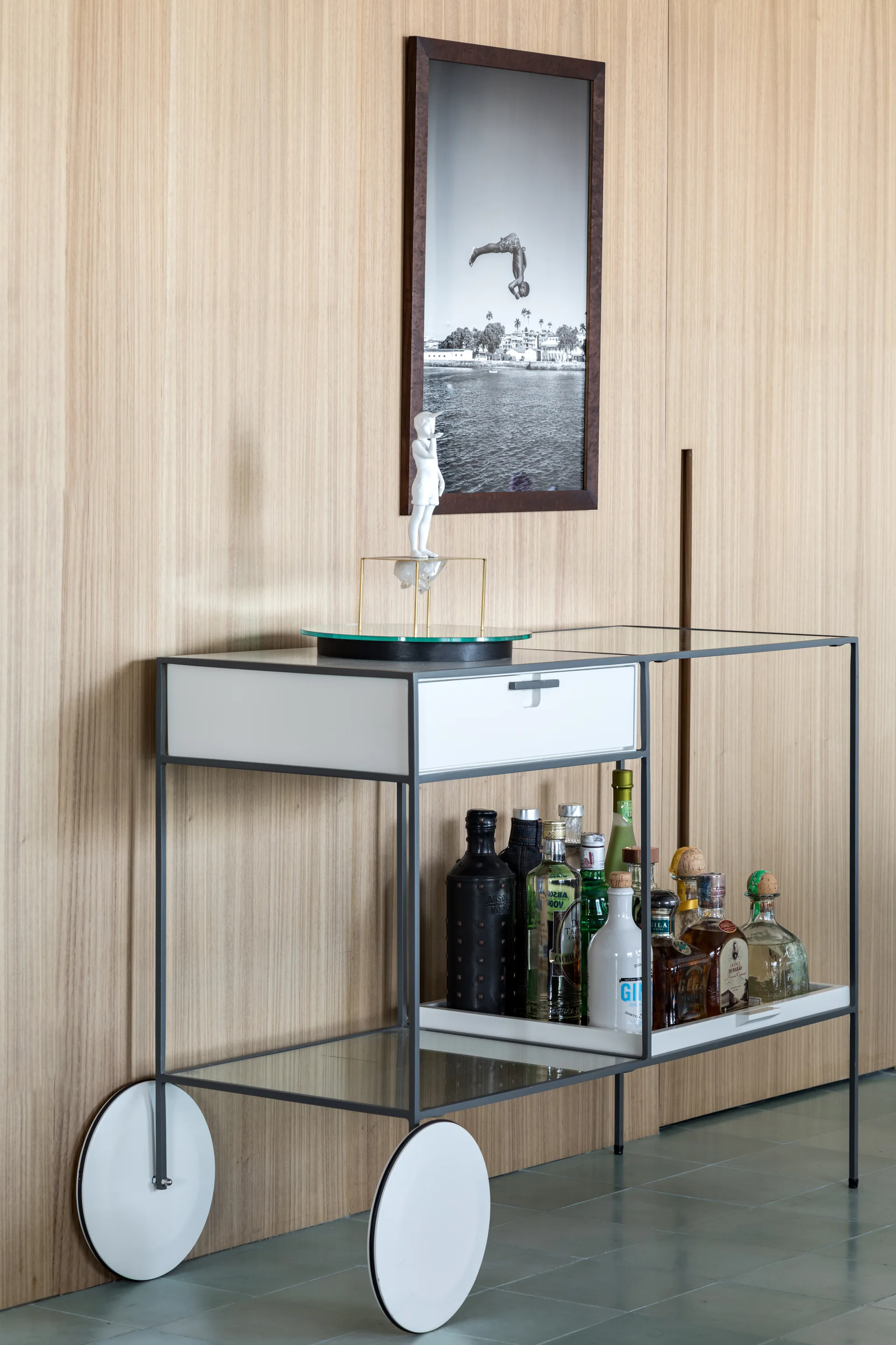 A modern metal-framed bar cart with glass shelves and large white wheels sits against a vertical wood-paneled wall displaying liquor bottles and art.