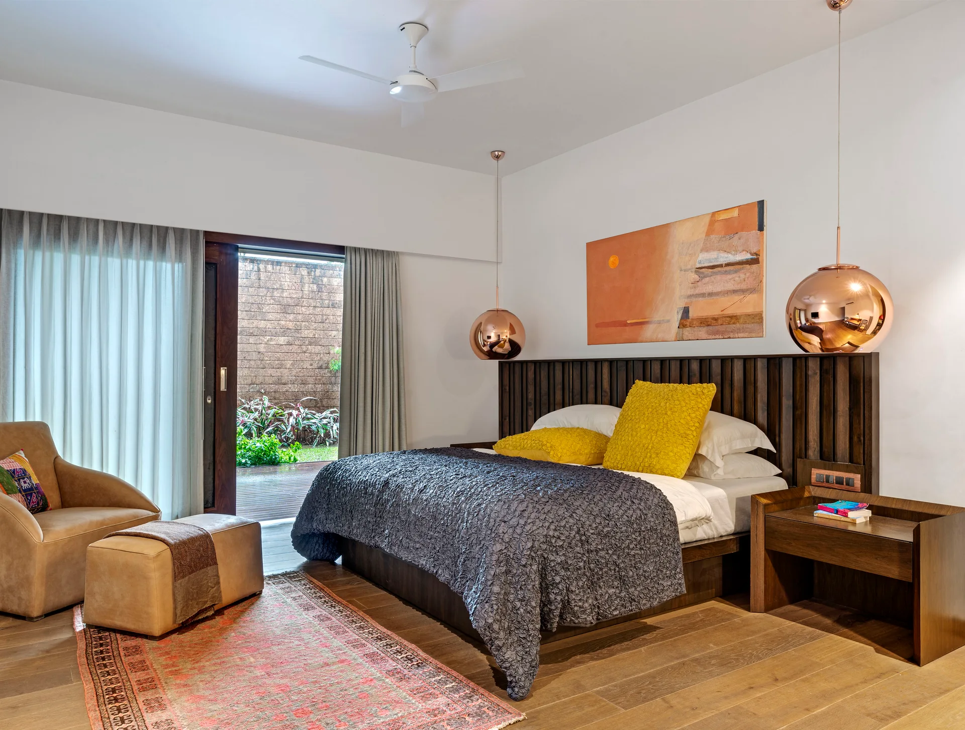 A contemporary bedroom features a dark wood slatted headboard, two copper pendant lights, a textured grey bedspread, and a sliding glass door.