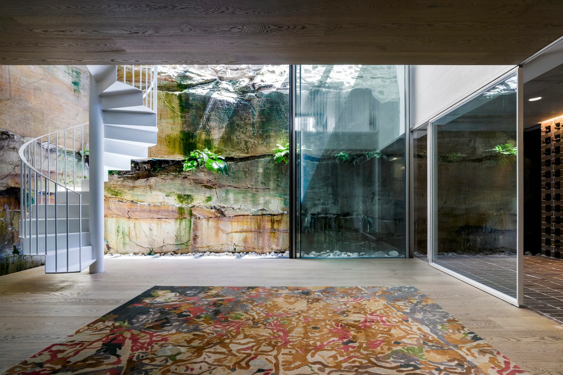 A white metal spiral staircase stands next to a floor-to-ceiling glass window showing a mossy natural rock wall.