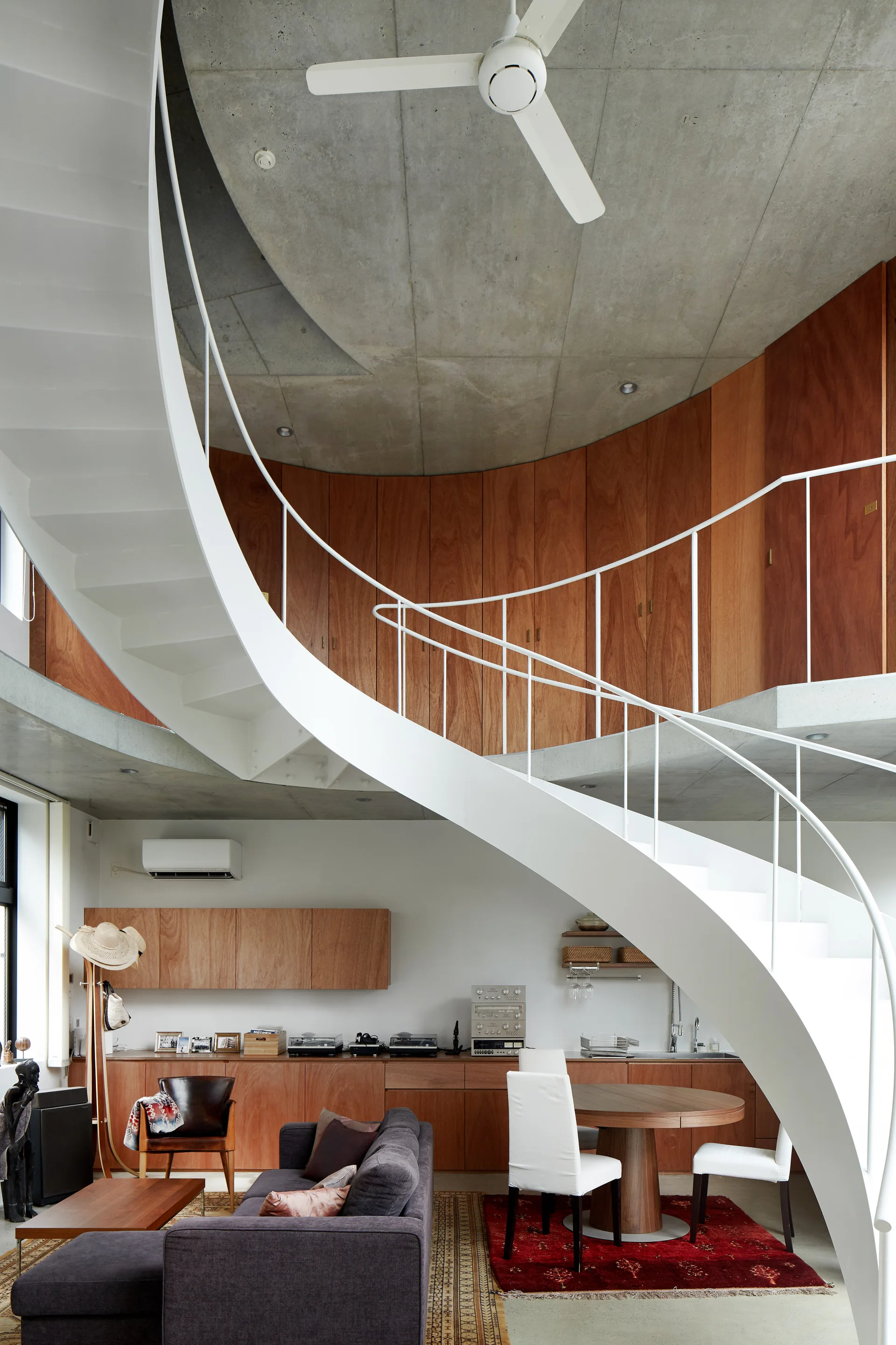 A white spiral staircase winds upward toward a concrete ceiling and curved wood-paneled walls in a modern open-concept living space.