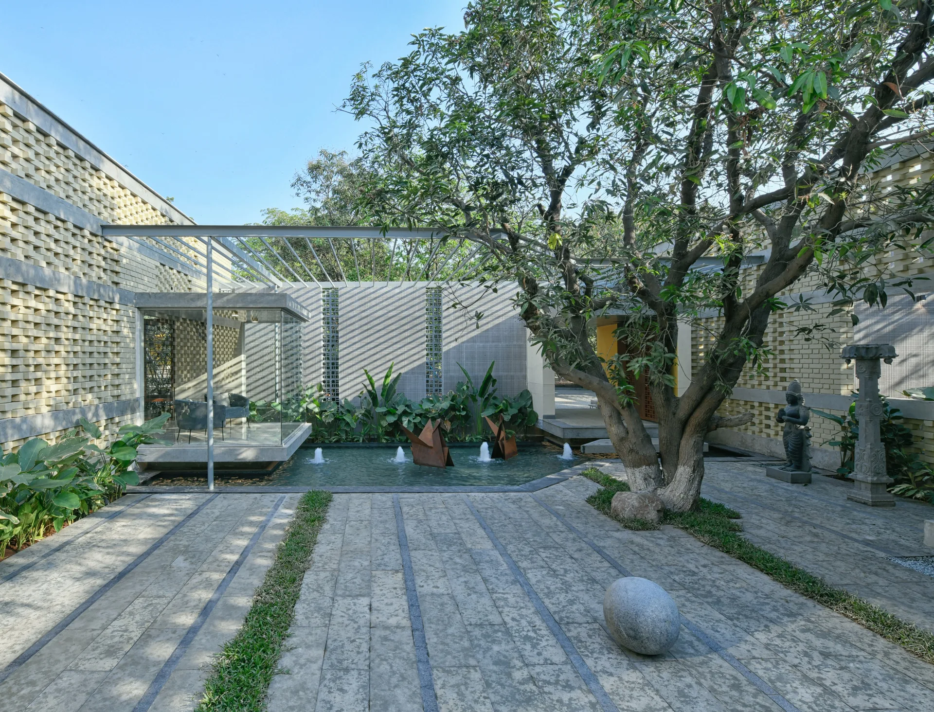 A modern outdoor courtyard featuring a stone-paved path, a mature tree, and a small pool of water with fountain jets against a textured brick wall.