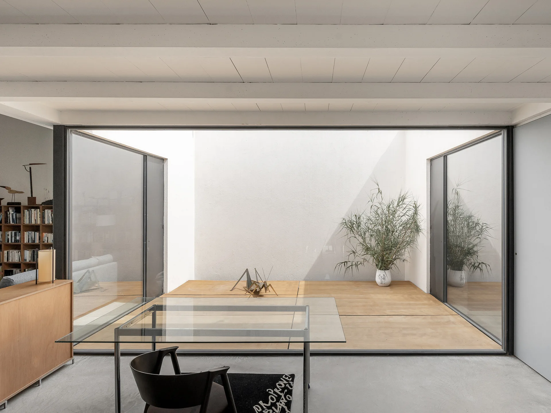 A glass dining table and black chair on a concrete floor look out toward an outdoor wooden courtyard through sliding glass doors.
