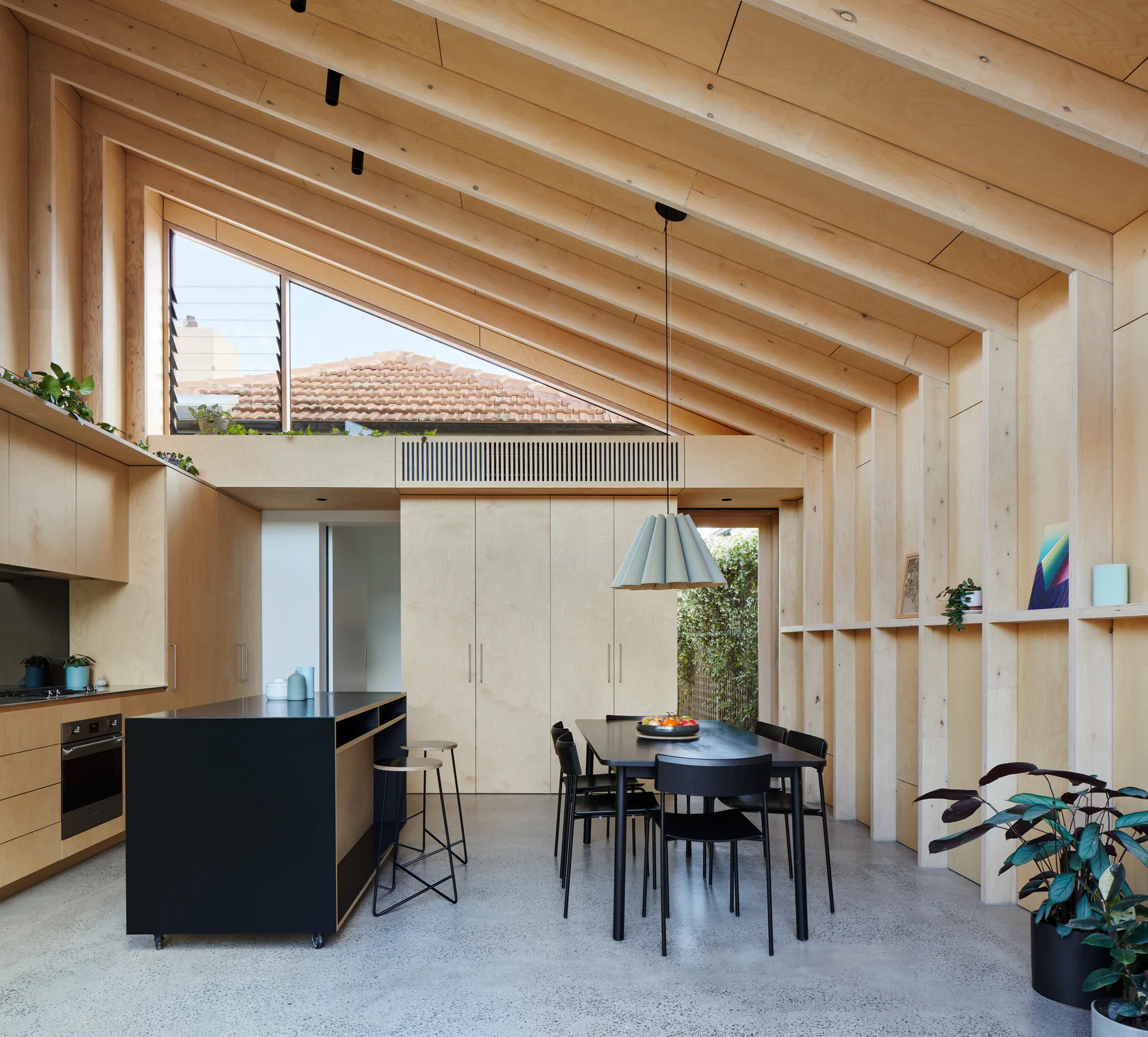 A modern open-plan kitchen and dining area featuring plywood paneled walls, a sloped ceiling, polished concrete floors, and black furniture.