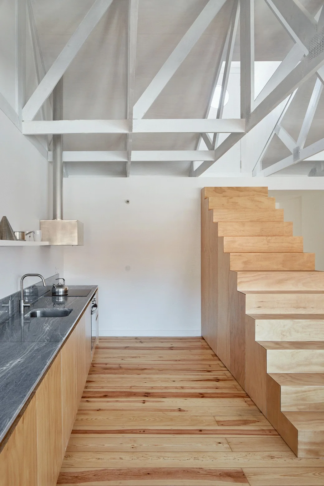 A minimalist kitchen features light wood cabinets, a dark grey countertop, and a matching plywood staircase beneath white exposed roof trusses.