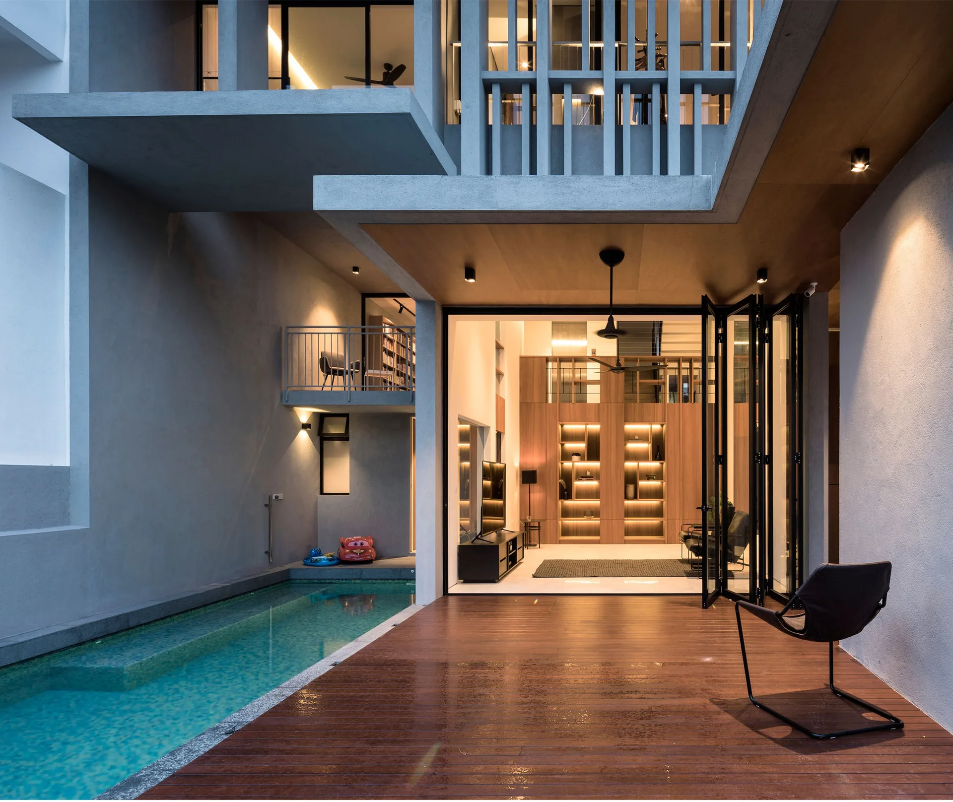 A modern home patio featuring a wooden deck next to a narrow lap pool and folding glass doors that open to an illuminated interior.