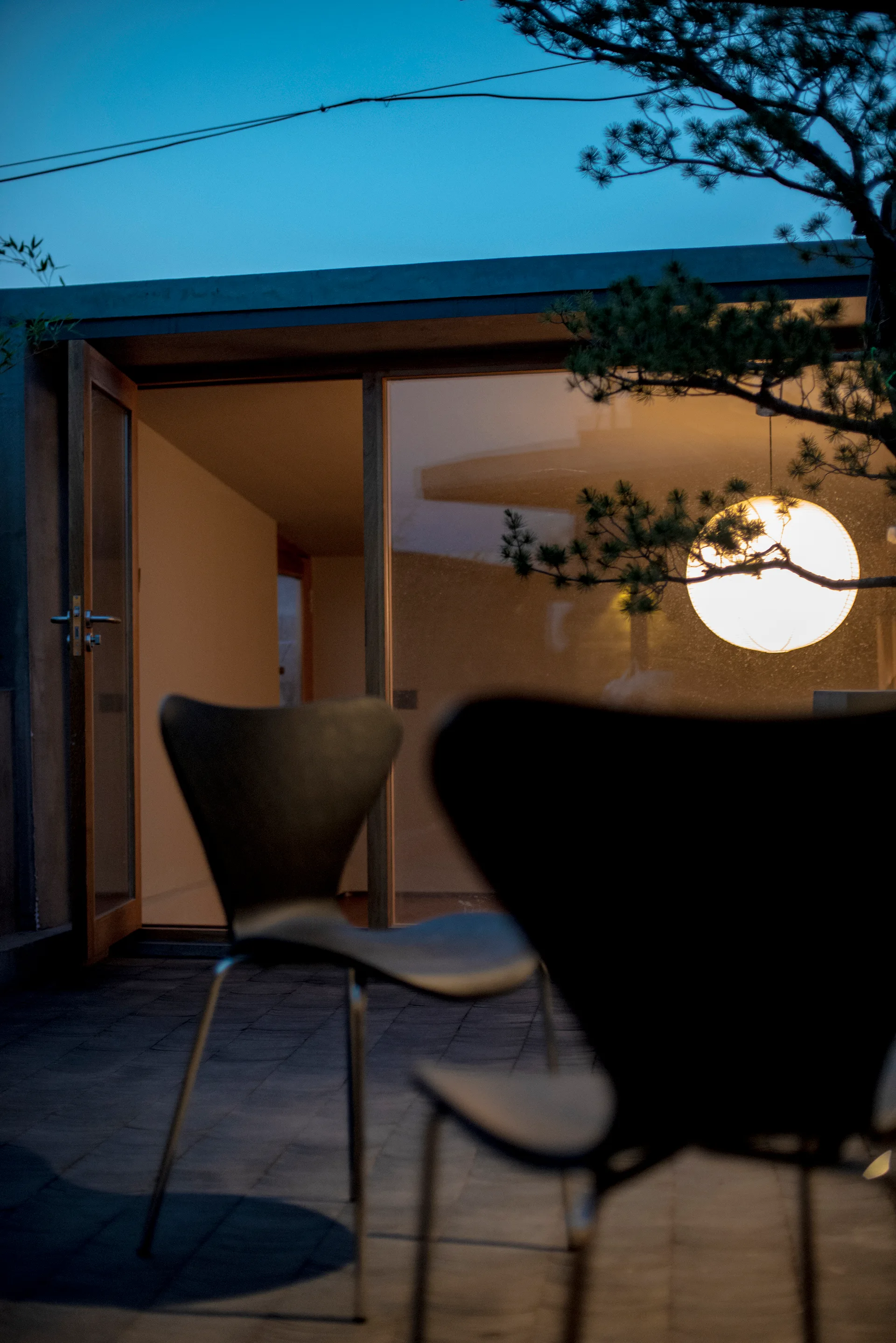 Two dark curved chairs sit on a paved stone patio at dusk looking towards an interior with a glowing round pendant lamp.