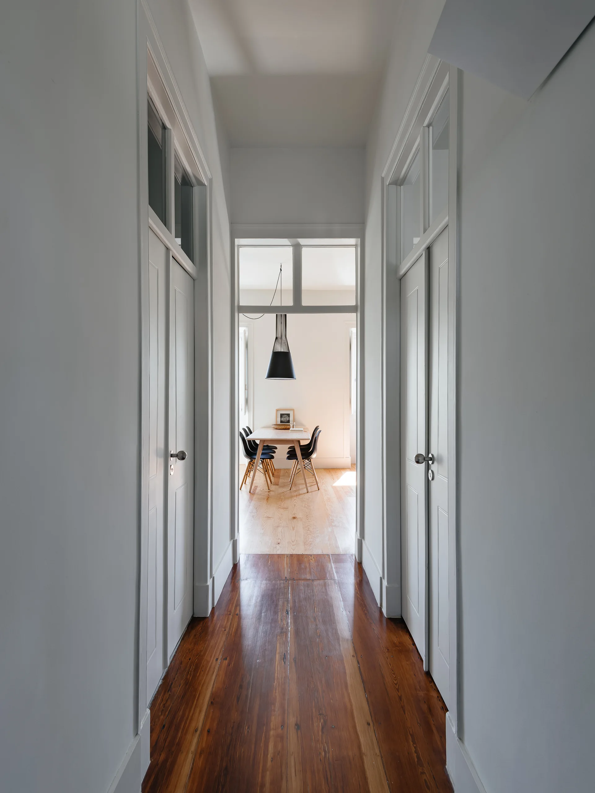 A narrow white hallway with paneled doors and glass transom windows leads to a brightly lit dining room with a wooden table and black chairs.