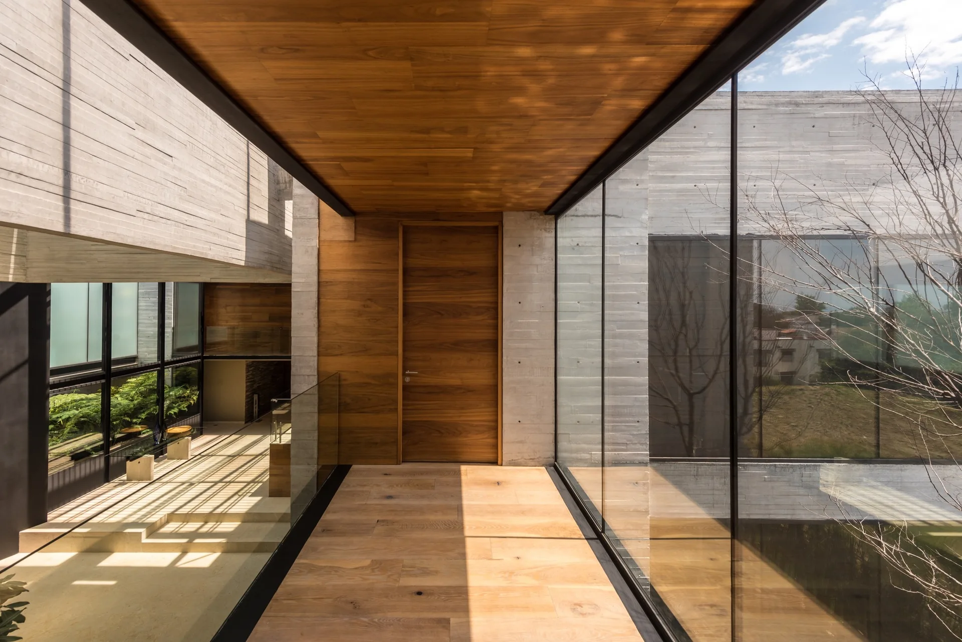 A minimalist hallway features warm wood flooring and ceiling, board-formed concrete walls, and large glass panels overlooking an exterior courtyard.