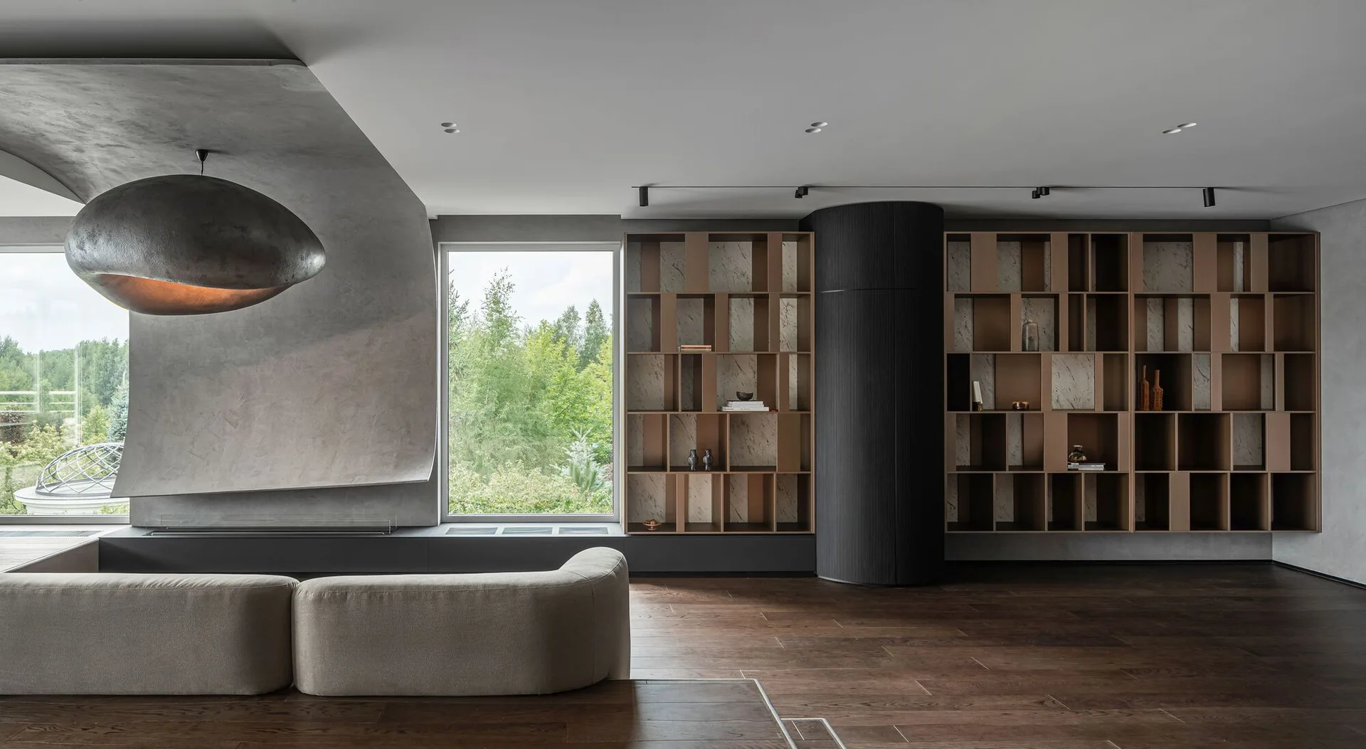 A minimalist living room with a curved concrete wall, a low beige sofa, dark wood floors, and open wooden shelving split by a ribbed black column.