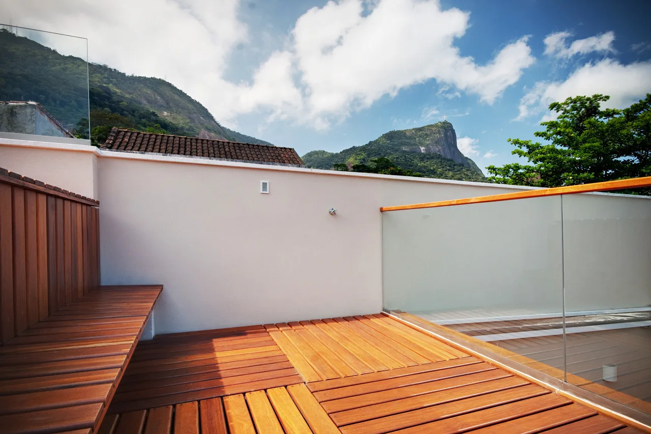 A wooden roof terrace features a built-in bench and a glass railing overlooking green mountains under a bright blue sky.