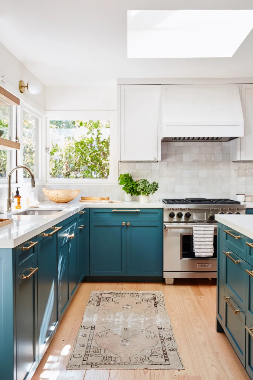 A bright kitchen features teal lower cabinets, white upper cabinets, a stainless steel range under a skylight, and light wood flooring with a runner rug.