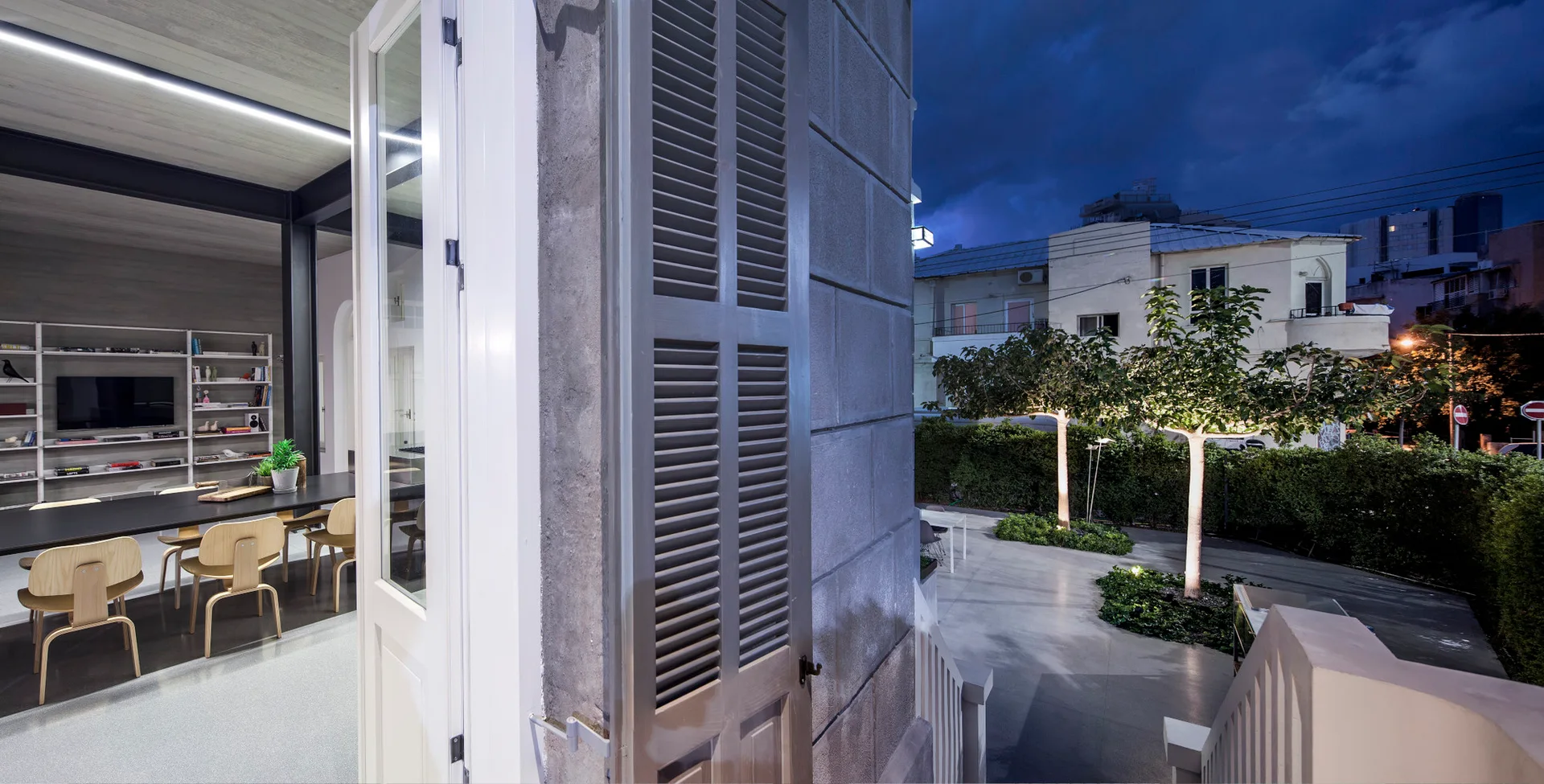 An open white doorway with grey slatted shutters connects a modern indoor dining area to an illuminated outdoor courtyard with green trees at night.