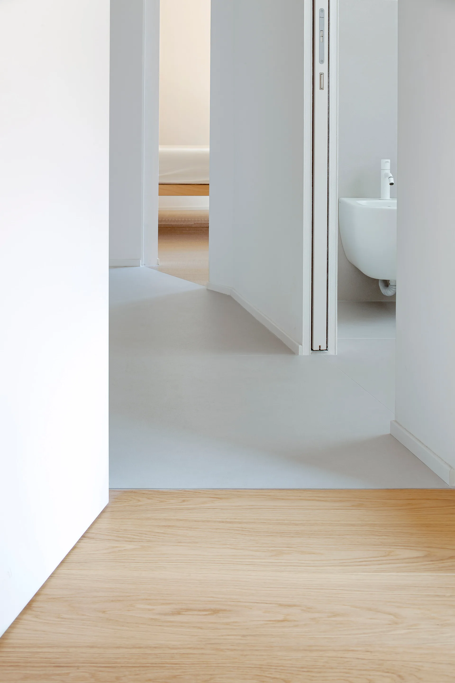 A minimalist hallway features a transition from light wood flooring to gray tile, with open doorways leading to a bedroom and bathroom.